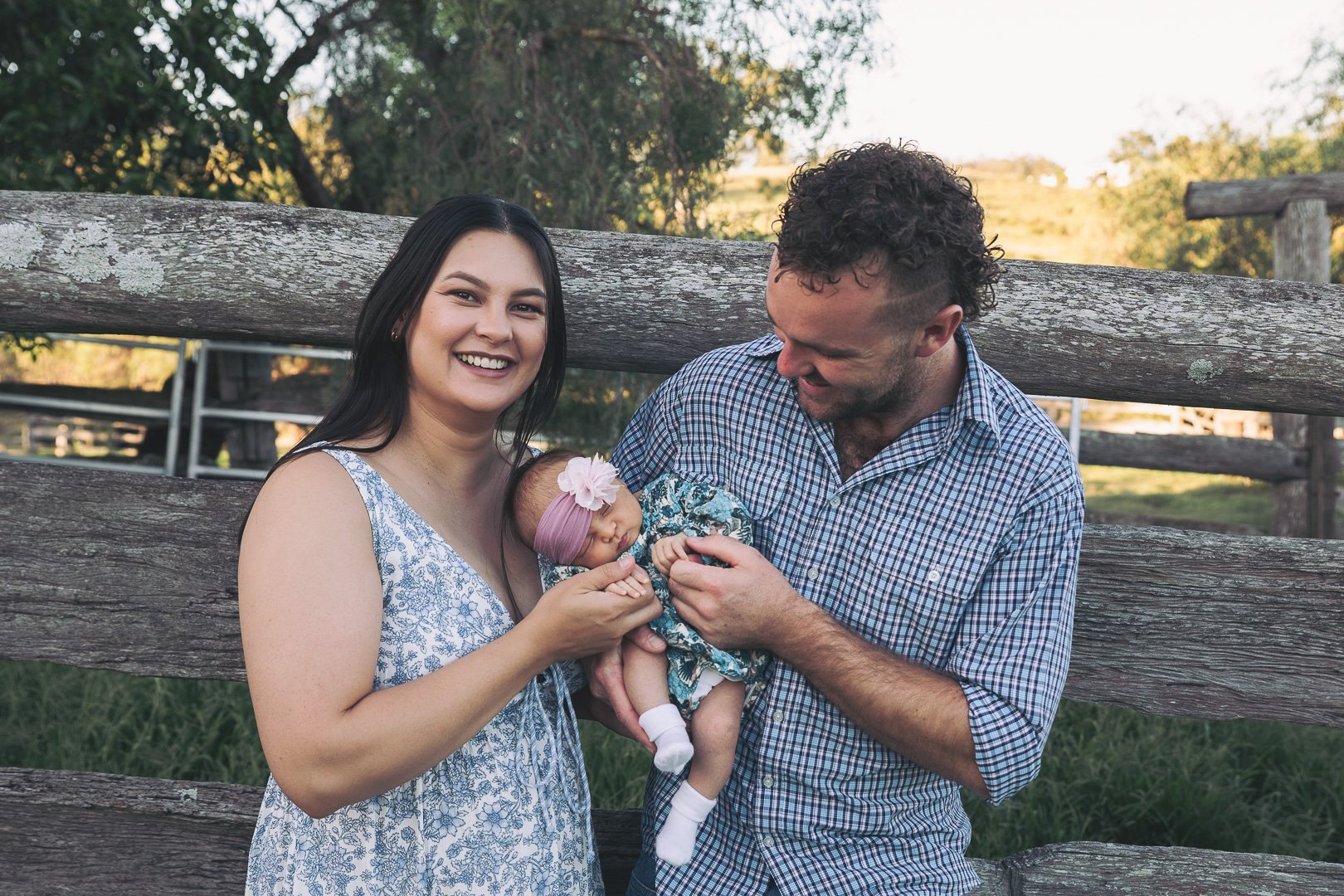 A happy family outdoors, with a woman, man, and a newborn baby, standing in front of a wooden fence, surrounded by trees and greenery.