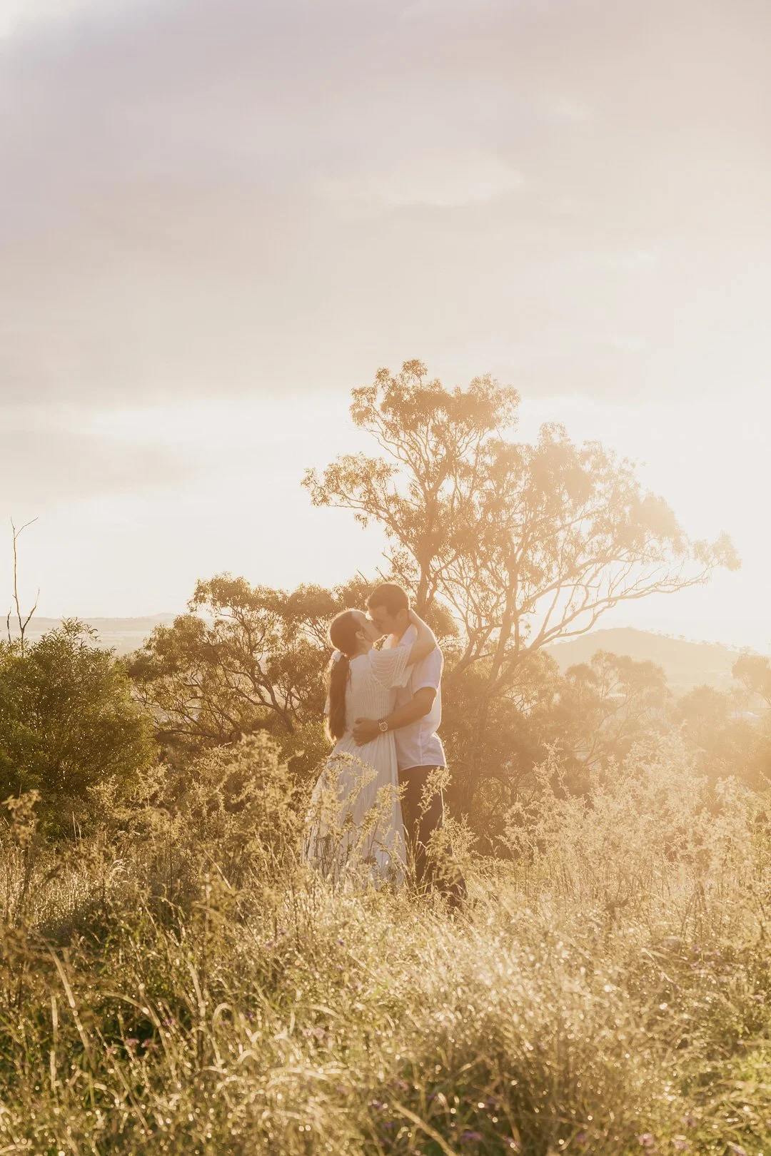 A couple embracing in a field at sunset with trees in the background.