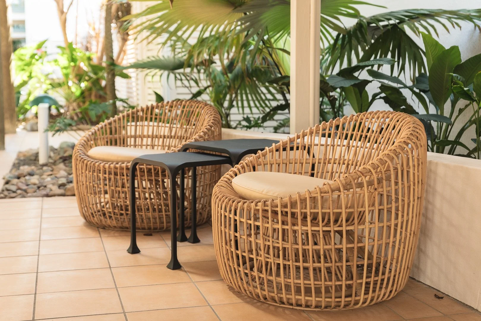 Two rattan chairs with cream cushions, two small black side tables, and lush tropical plants in the background, on a tiled outdoor patio. Styling photography, Laurene Bardsley Photography, Toowoomba Photographer