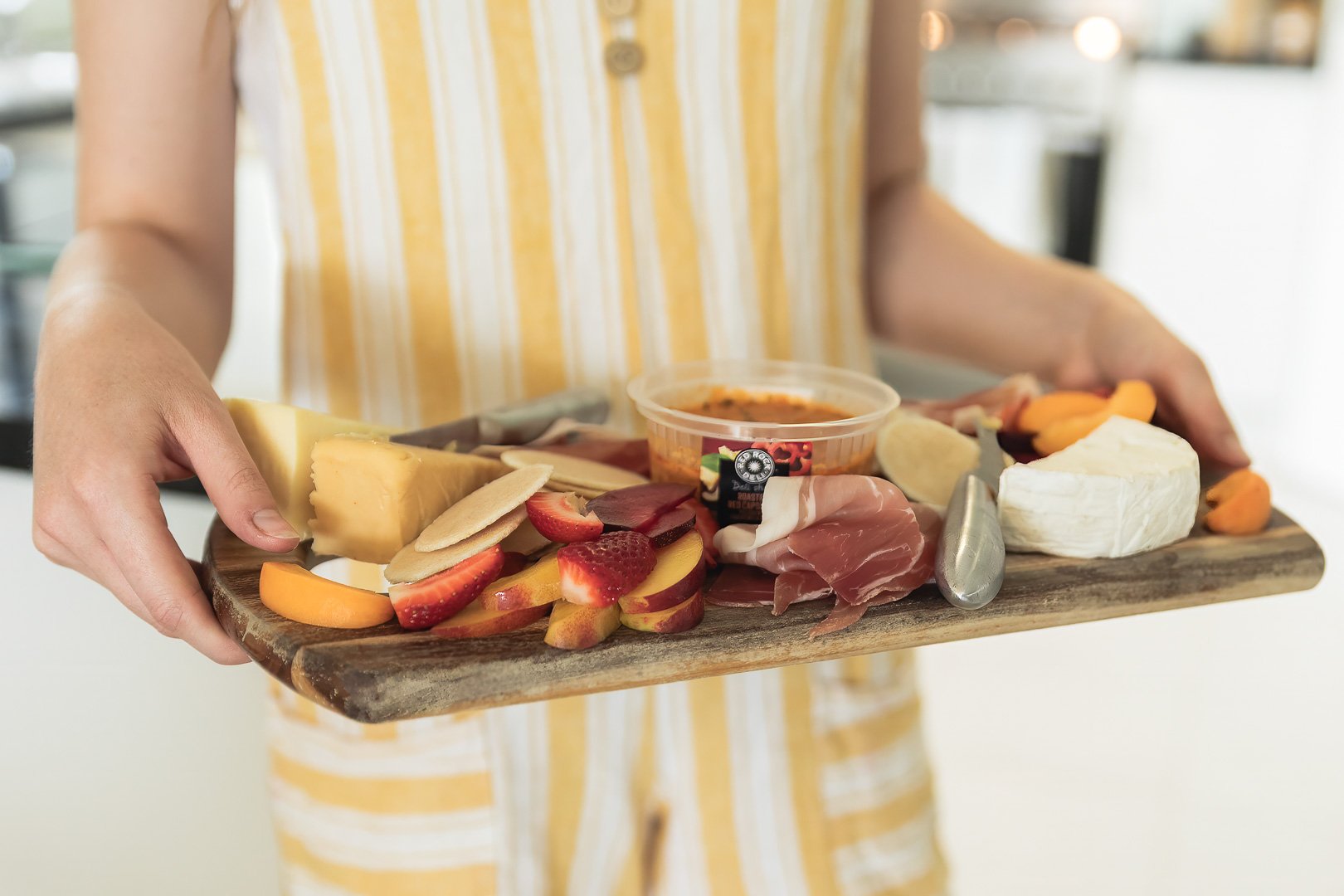 Person holding a wooden cheese and charcuterie platter with cheeses, strawberries, sliced peaches, prosciutto, and a small container of dipping sauce.
