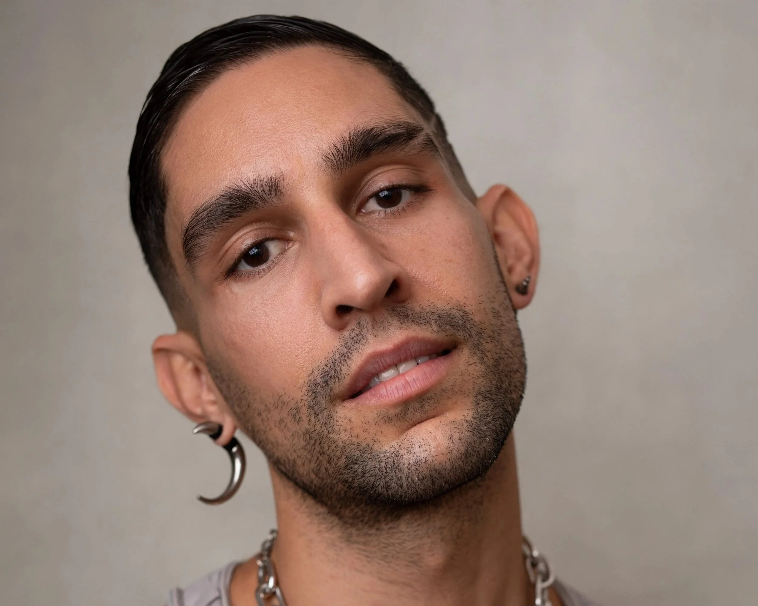 Close-up portrait of a young man with dark hair slicked back, wearing earrings and a chain necklace, with a neutral expression and a slight tilt of his head.