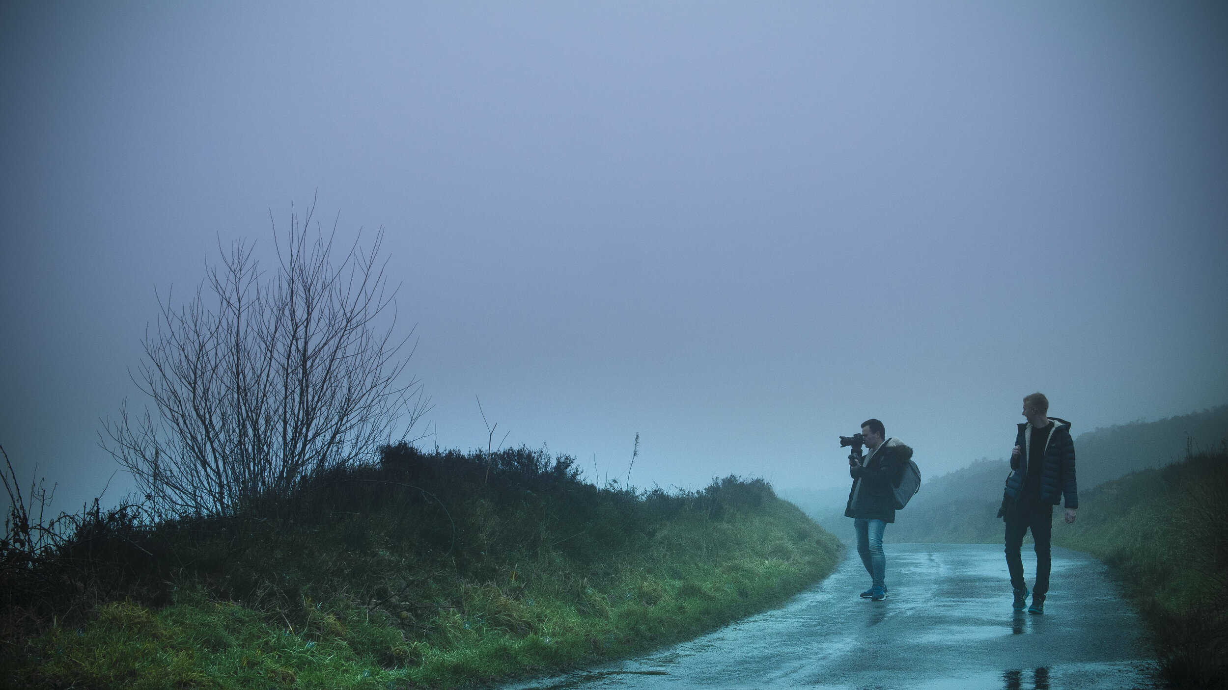 Two people walking on a foggy rural road, one holding a camera, with bare trees and grass on the roadside.