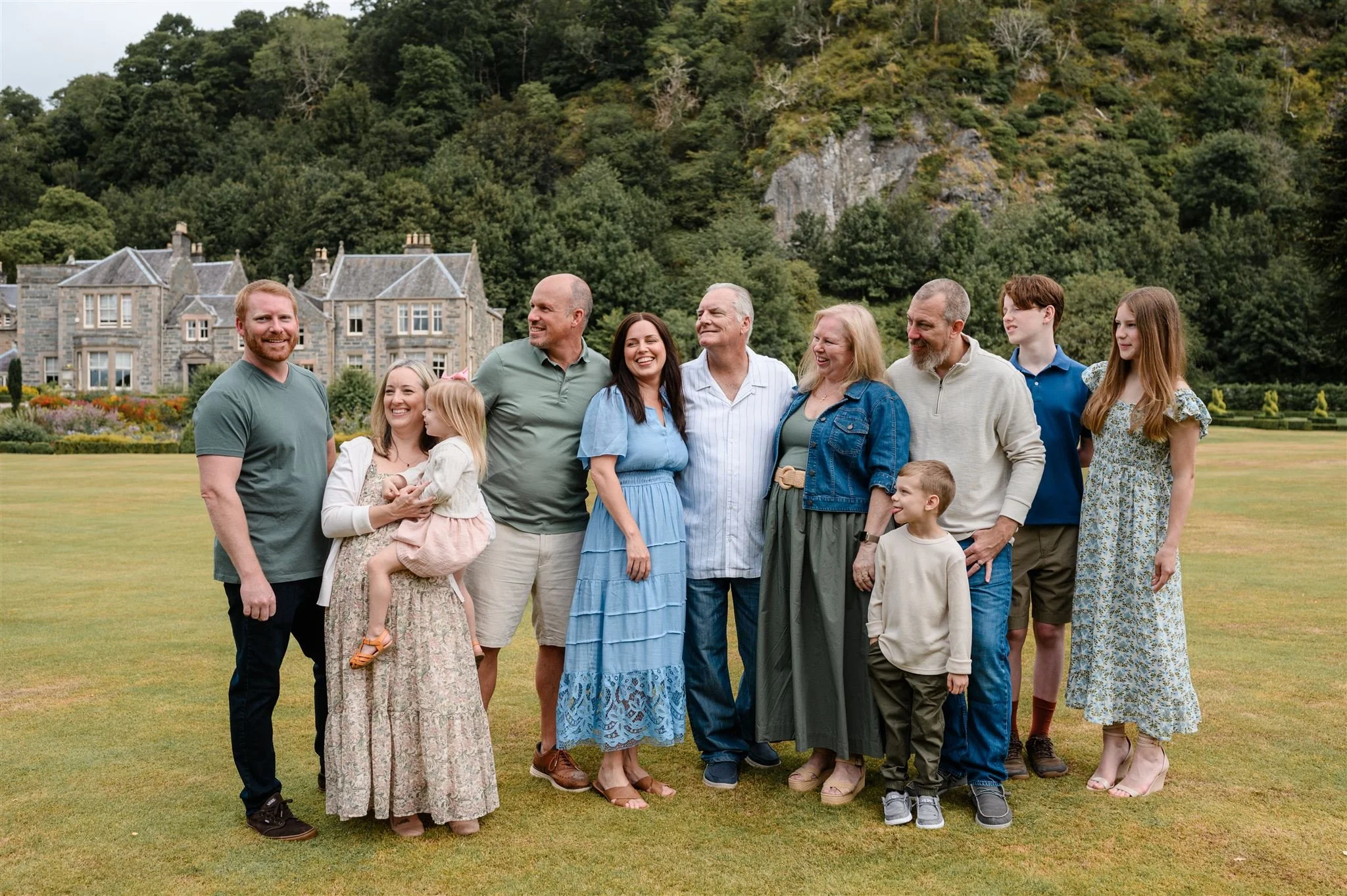 A family gathering outdoors on a lawn with a large house and wooded hillside in the background. The group includes adults and children dressed in casual summer clothing, smiling and standing close together.