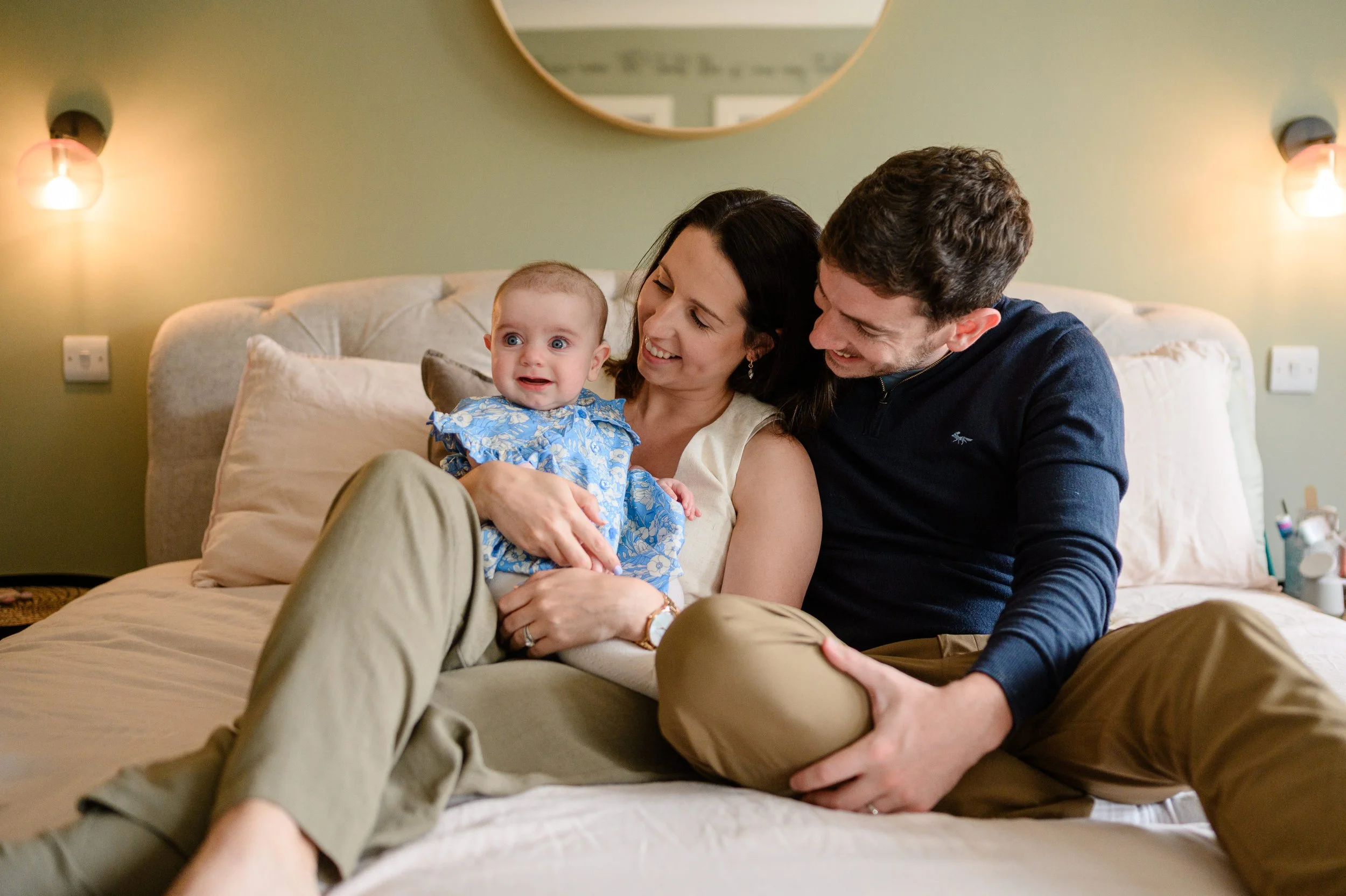 A family of three sitting on a bed, smiling and playing with a baby girl who is wearing a blue floral dress.