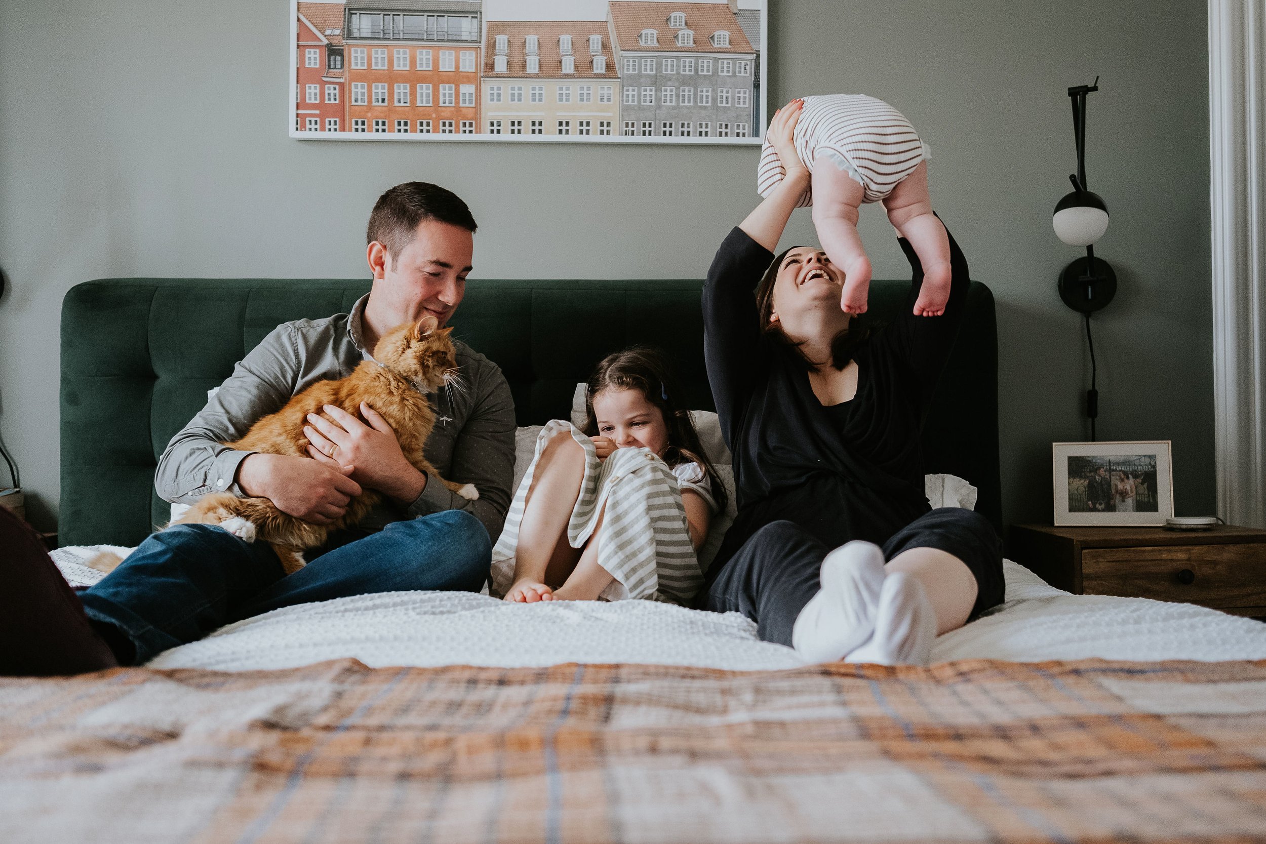 A family of four and a cat sitting on a bed, with a woman lifting a baby in the air, smiling. The man is holding the cat, and a young girl is lying next to him, also smiling. The room has gray walls, colorful cityscape artwork, a lamp, and a framed photo on a nightstand.