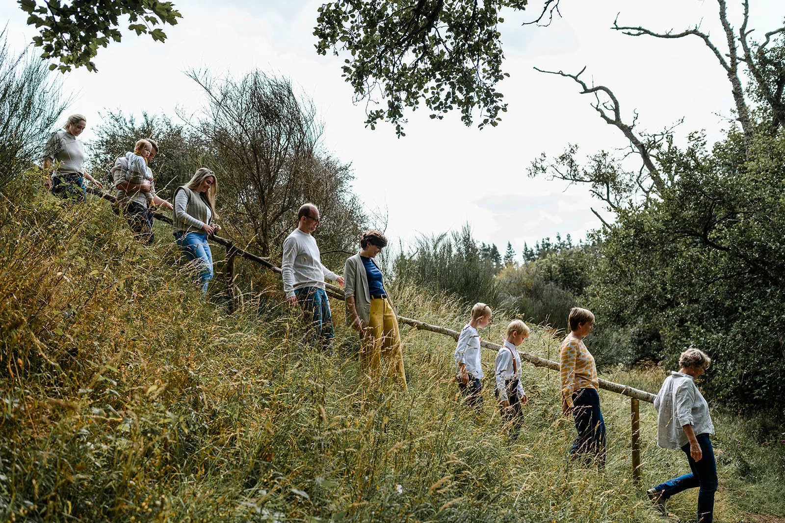 A group of people descending a grassy hillside along a wooden rail fence on a cloudy day, surrounded by trees and bushes.