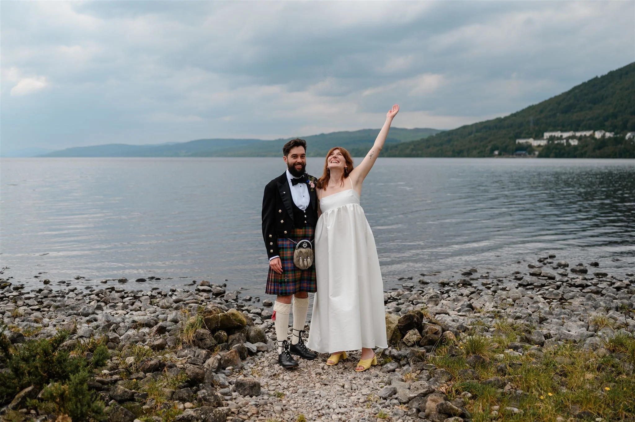 A couple celebrating their wedding by a lake, with the bride raising her arm and the groom in a kilt, both smiling.