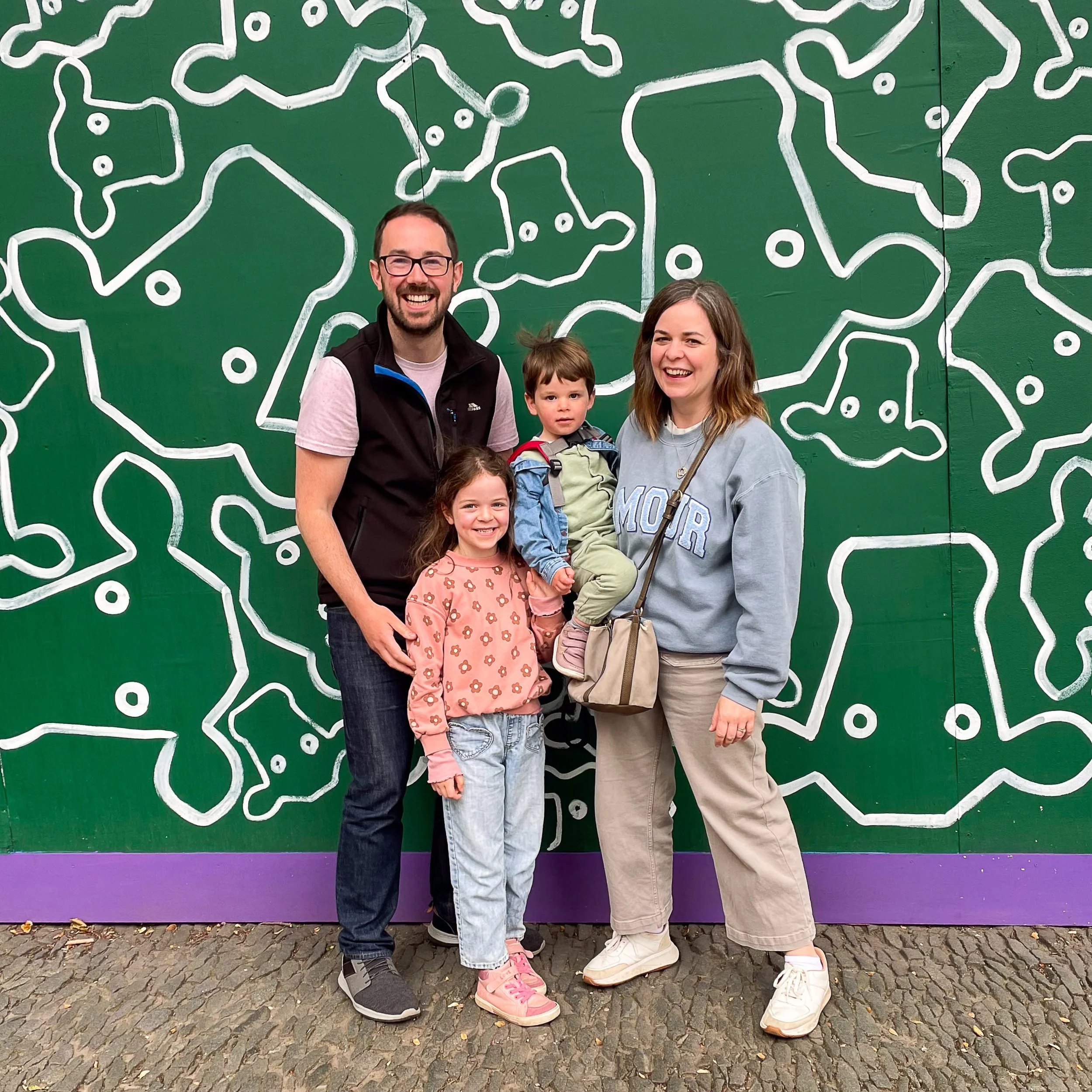 A family of four standing in front of a green wall. The father has glasses and a beard, wearing a black vest over a pink shirt. The mother has shoulder-length hair, wearing a gray sweatshirt with the word 'MOM' on it. The son, carried by the mother, has short hair and a backpack. The daughter, standing next to her father, has long hair and is smiling.