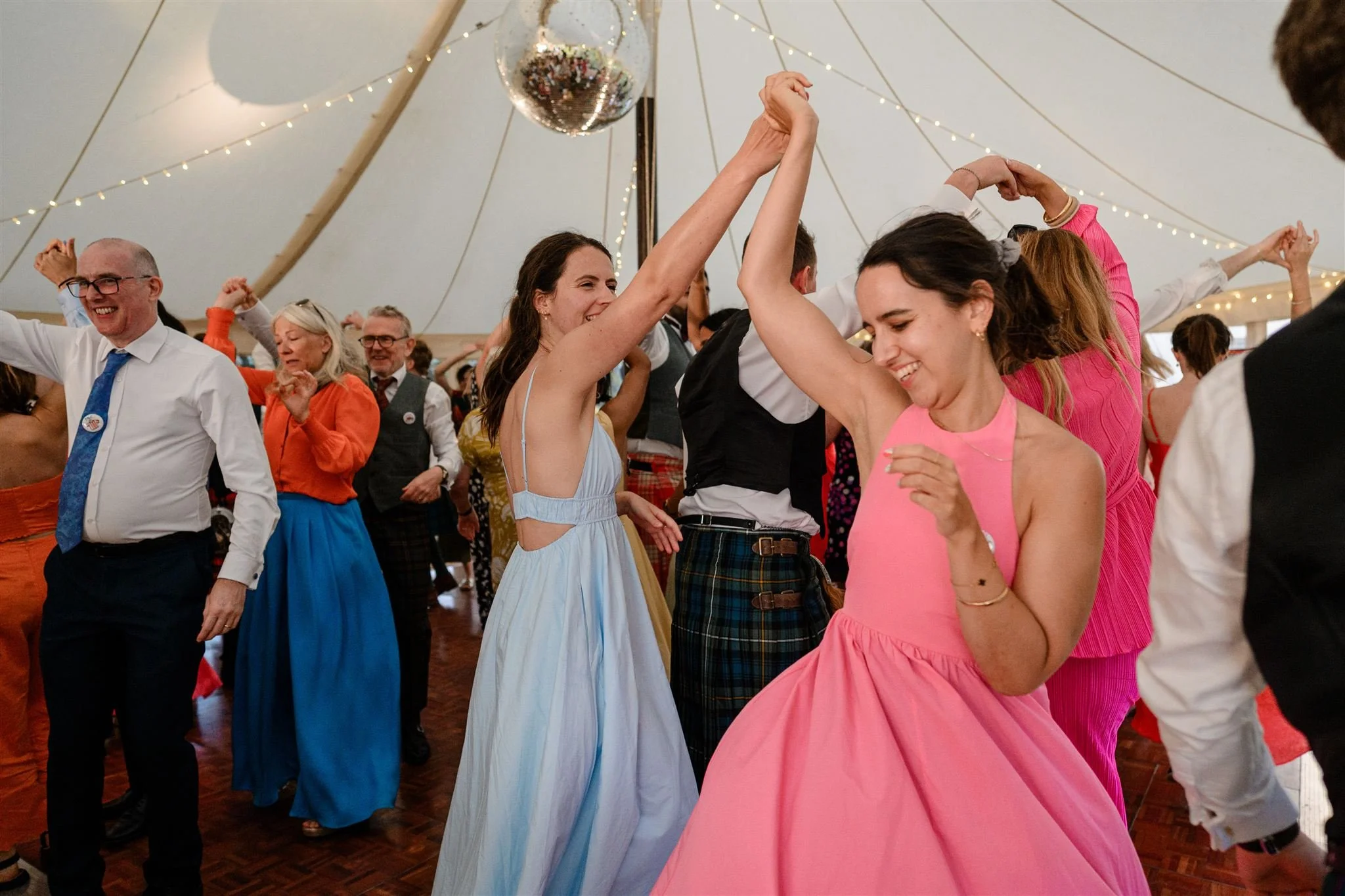 People dancing at a wedding reception under a decorated tent, with string lights and a disco ball overhead.