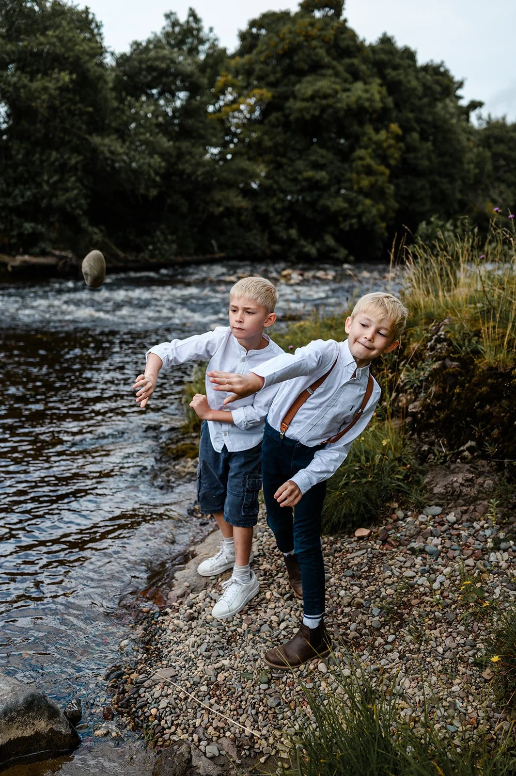 Two young boys playing by a river, with one throwing a stone into the water and the other watching, surrounded by trees and grass.