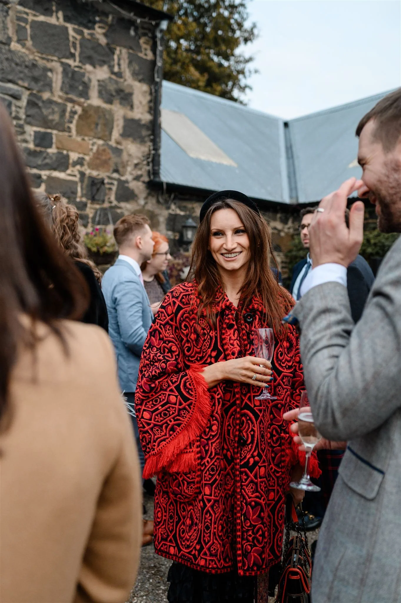 Group of people at an outdoor gathering, woman in a red patterned shawl holding a glass of champagne, smiling, talking to a man in a grey suit, brick building in the background.