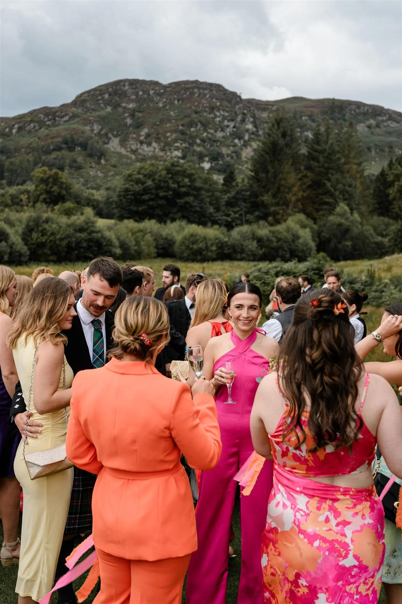 A group of people socializing outdoors, dressed in colorful formal attire, with a scenic mountain landscape in the background.