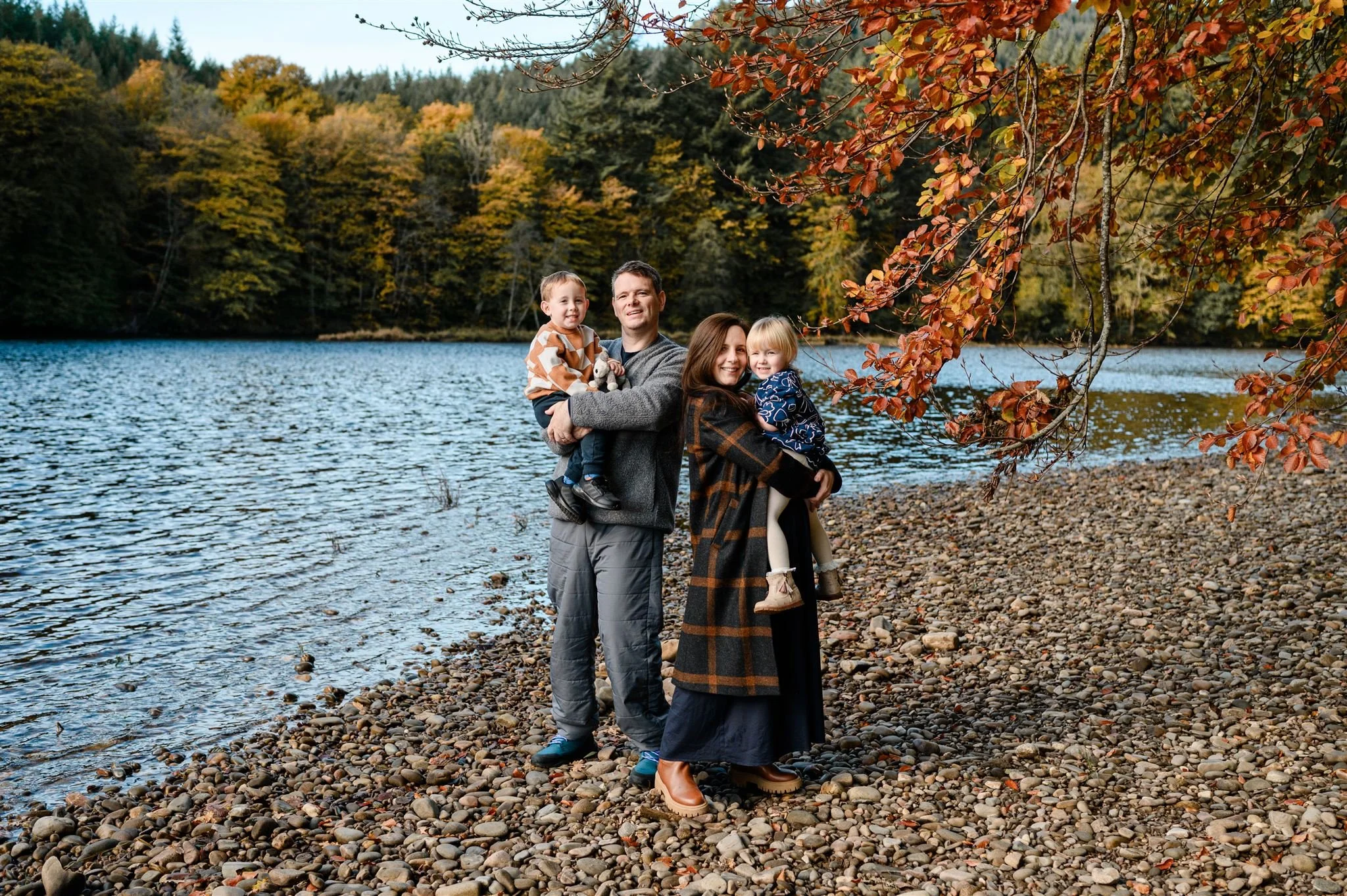 A family of four standing on a rocky shoreline by a lake in autumn. The father and older daughter are holding younger children, with trees with fall foliage in the background.
