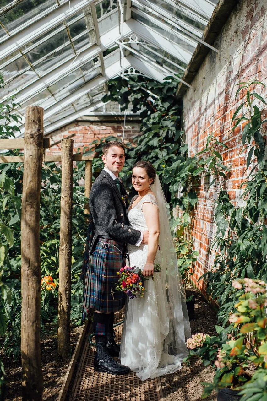 A bride and groom standing together inside a greenhouse at Errol Park with plants and brick walls, dressed in wedding attire. The bride is holding a colorful bouquet and smiling, while the groom is wearing a traditional kilt and blazer.