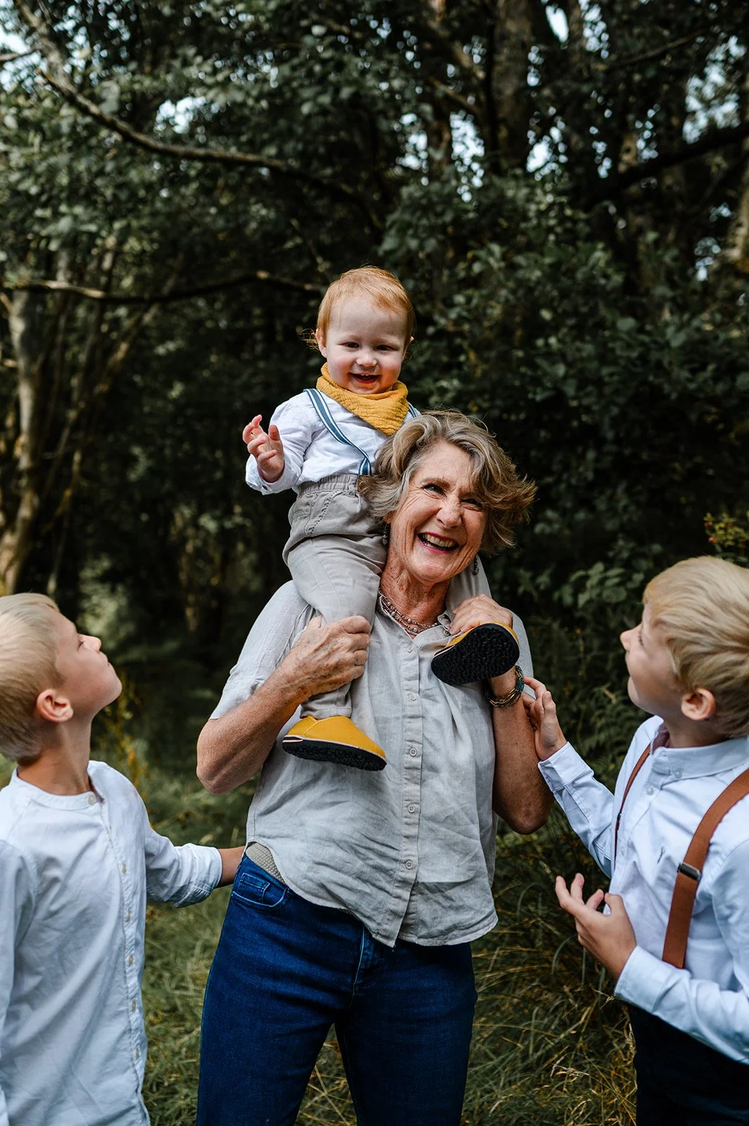 An elderly woman, smiling, holding a young child on her shoulders, surrounded by three young boys in an outdoor wooded area.