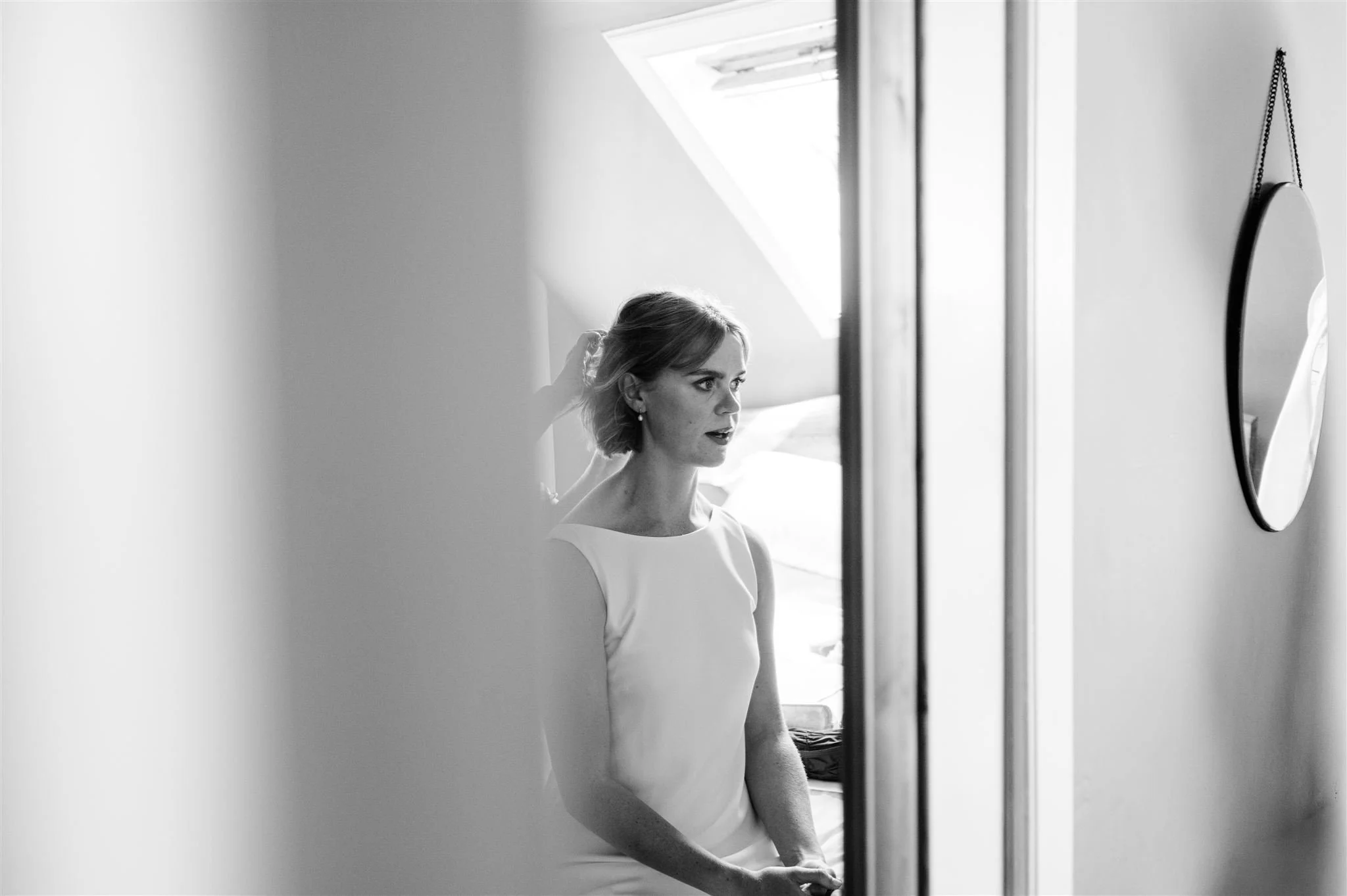 A woman in a white dress with a ribbon in her hair, looking into a mirror in a bright room with a sloped ceiling and a skylight.