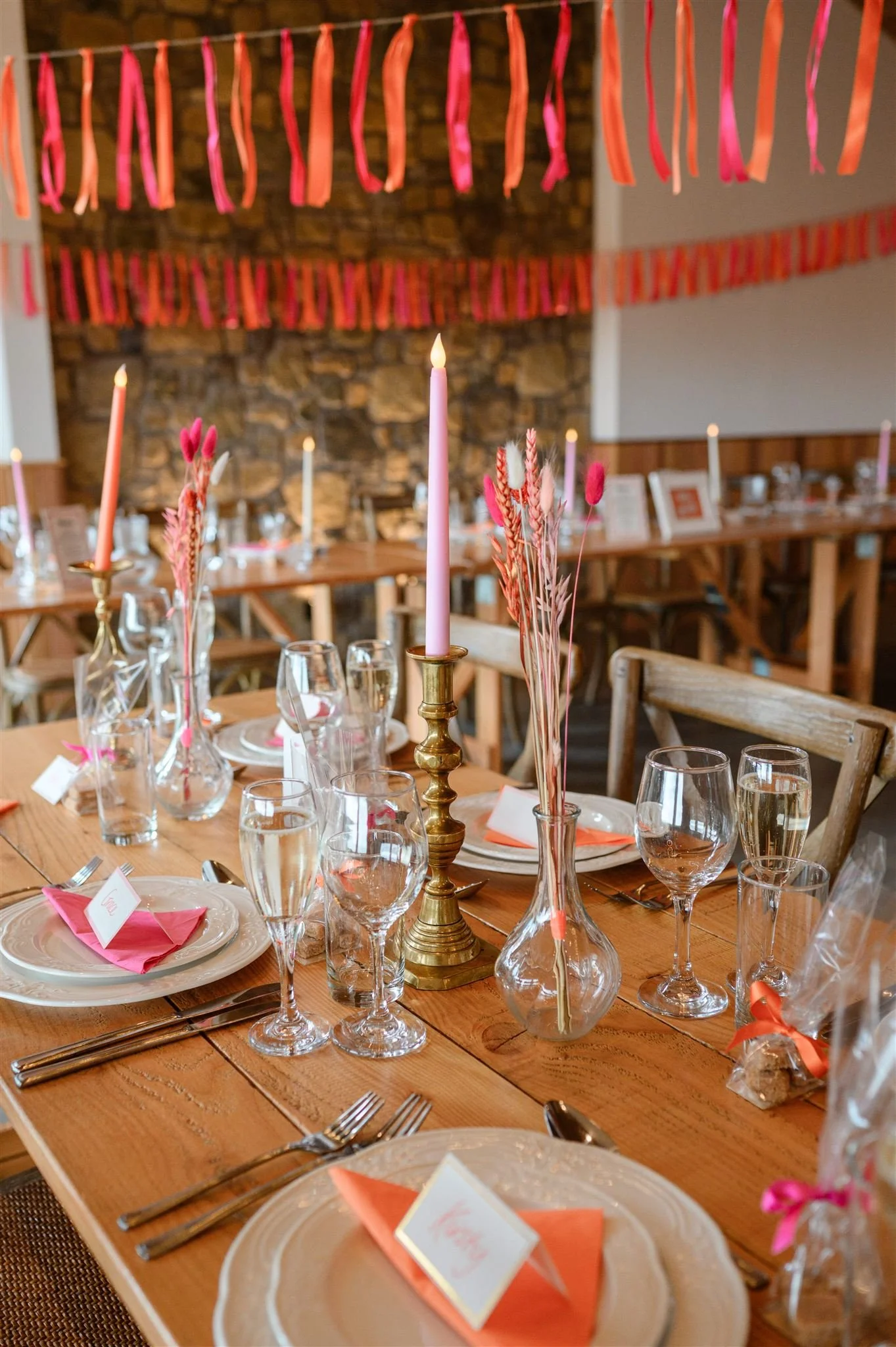 Decorated banquet table with pink candles, pink and orange paper napkins, glassware, and name cards, set for a celebration in a rustic event space with pink streamers hanging from the ceiling.