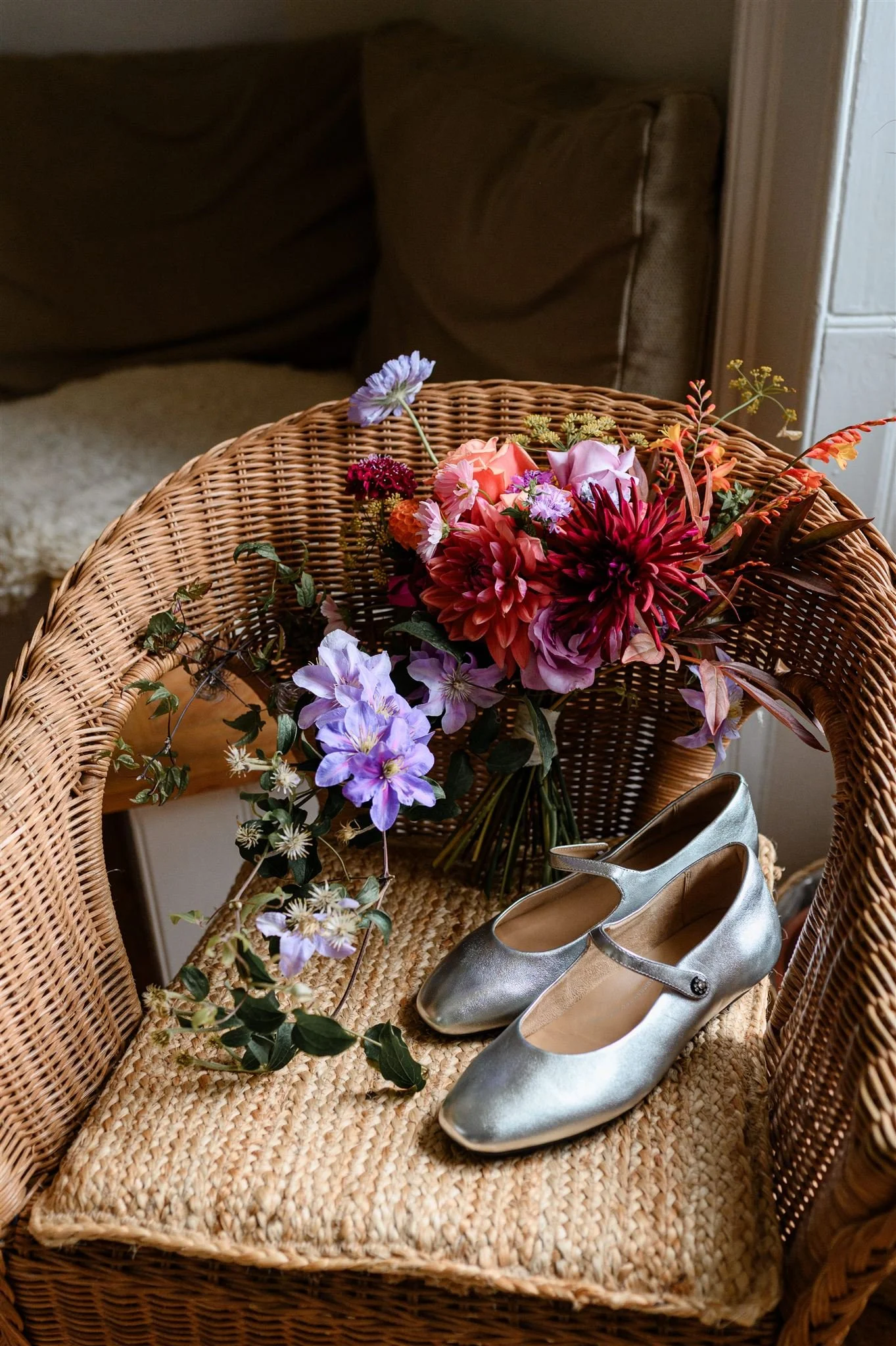Silver wedding shoes with a bouquet of colorful flowers on a wicker chair.