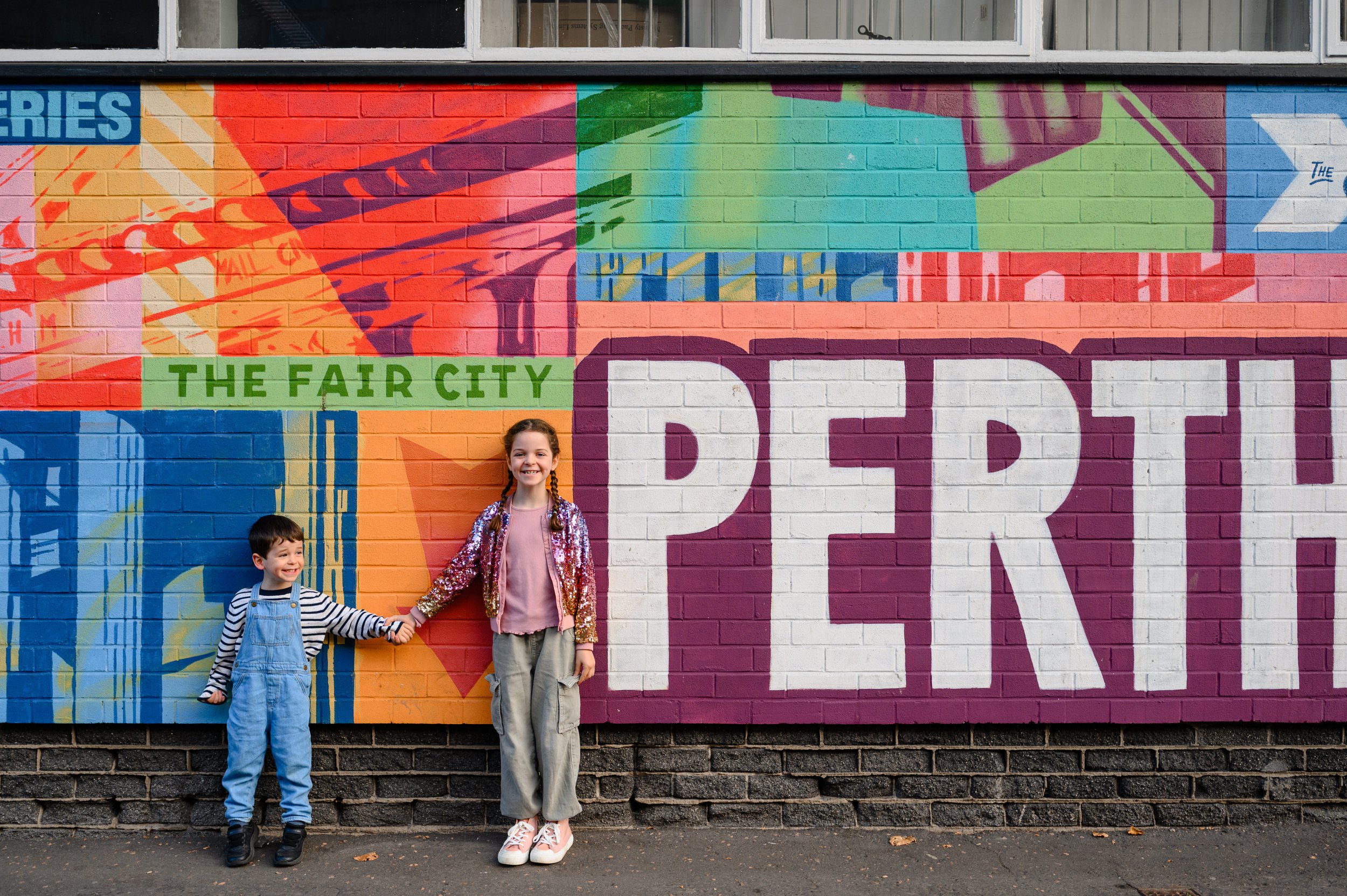 Two children, a boy and a girl, standing in front of a colorful mural on a brick wall. The mural reads 'The Fair City' and 'PERTH' with various abstract patterns and shapes.