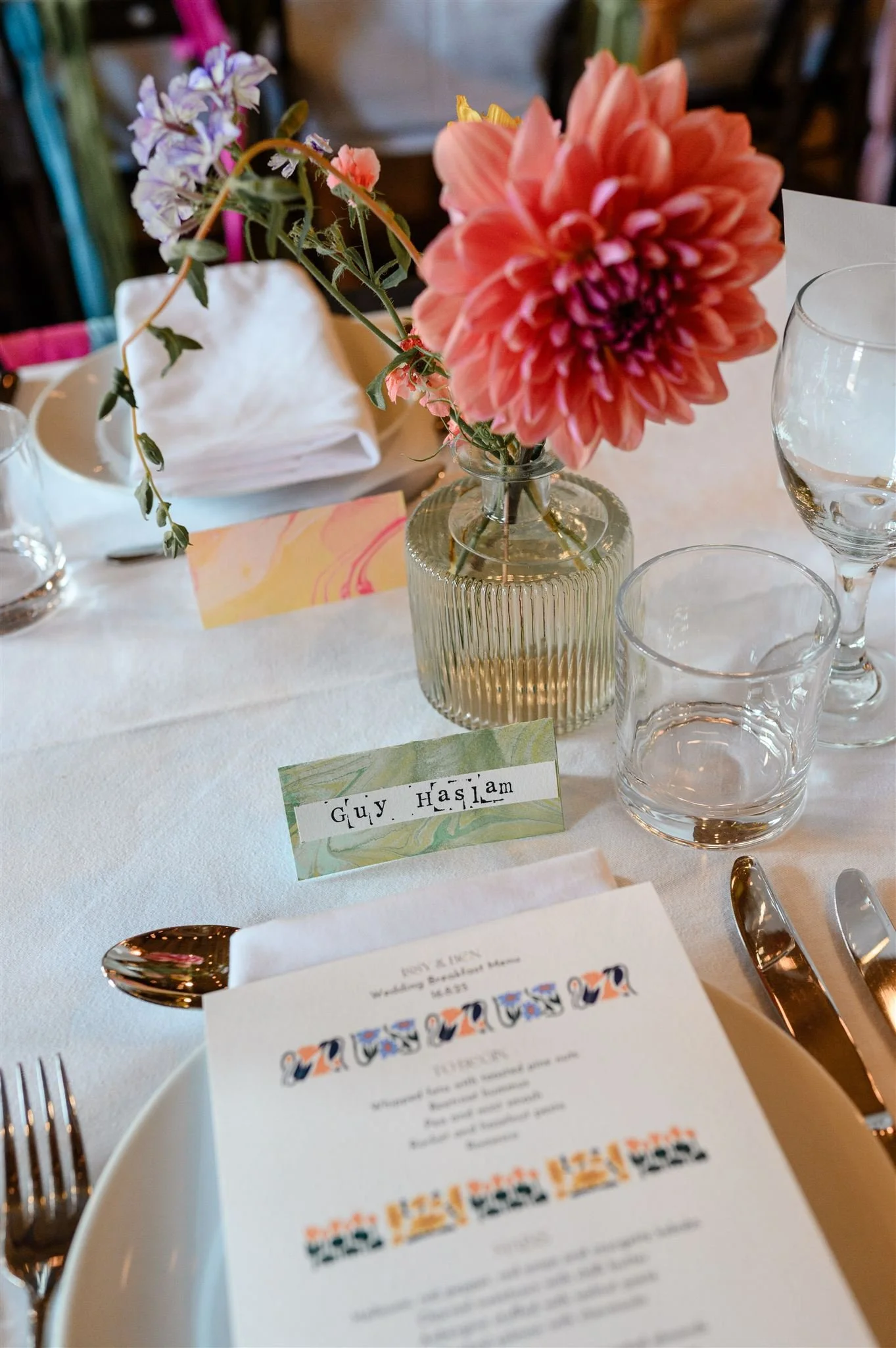 A table setting at a wedding reception with a floral centerpiece in a ribbed glass vase, a place card reading "Guy Haslam," a menu, and various glassware and cutlery arrangement.