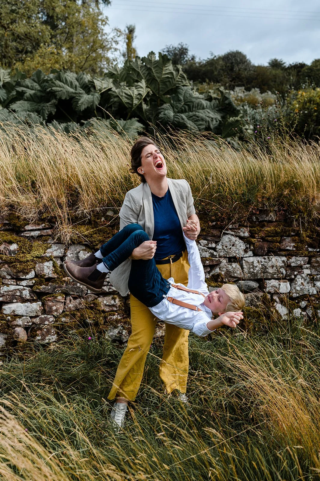 A woman with short dark hair in a gray jacket, and a young boy with blond hair in a white shirt and dark jeans, are outdoors amidst tall grass and large green plants. The woman appears distressed and is holding the boy who looks frightened and is rea