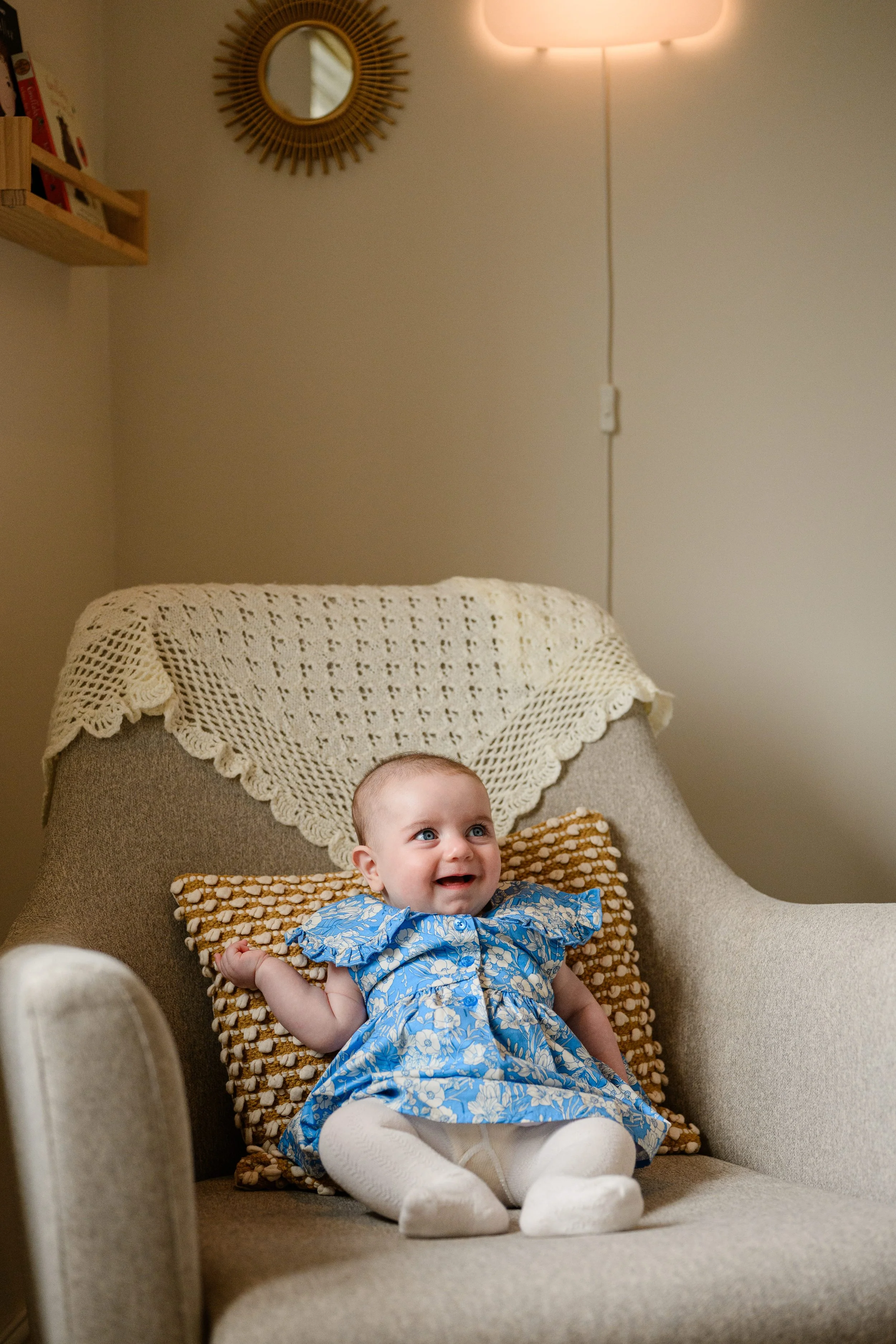 A smiling baby girl with blue eyes, dressed in a blue floral dress and white tights, sitting on a beige armchair with a textured pillow, in a cozy living room with a wall mirror and a wall-mounted light fixture.