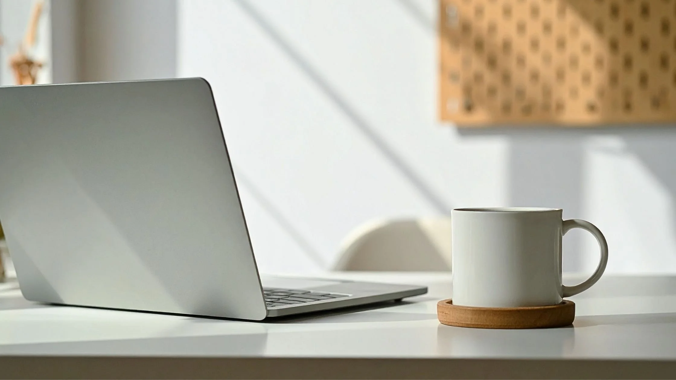 Minimalist desk setup with an open silver laptop and a white coffee mug in soft sunlight.
