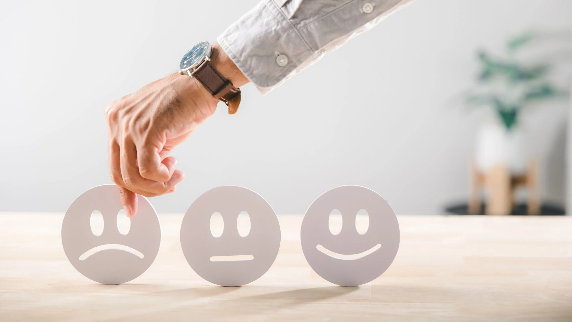 A hand wearing a wristwatch picks up a sad face icon next to neutral and happy face icons on a wooden table, symbolizing a website diagnostic audit or customer feedback review.