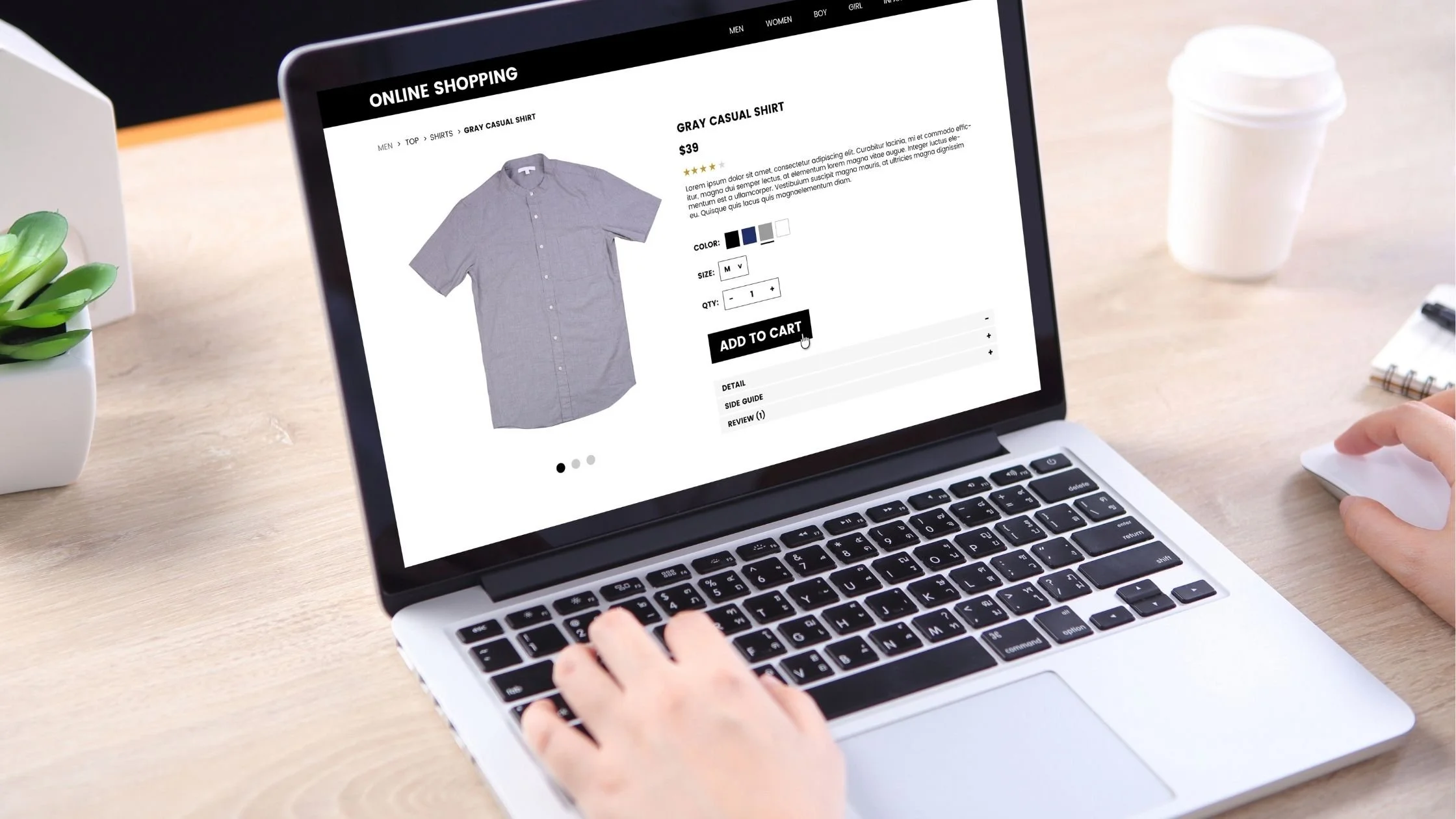 Person shopping online for a gray casual shirt on a laptop with an e-commerce website open on a wooden desk.