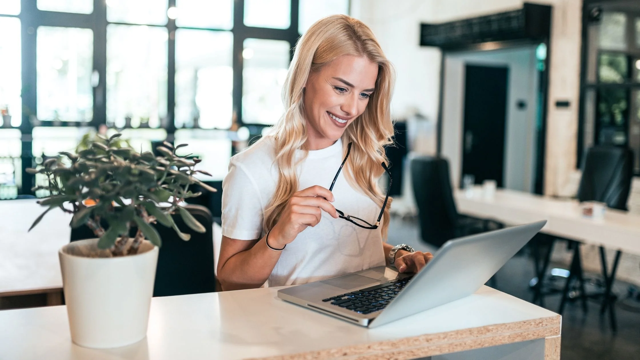 Smiling woman working on a laptop in a modern office space while holding eyeglasses and looking at the screen.
