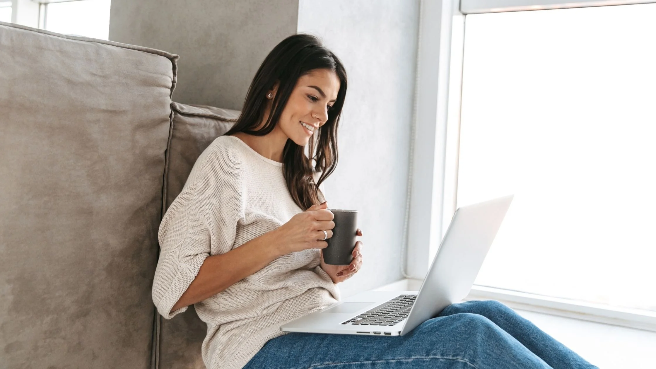 A smiling woman sitting by a bright window using a laptop while holding a mug.