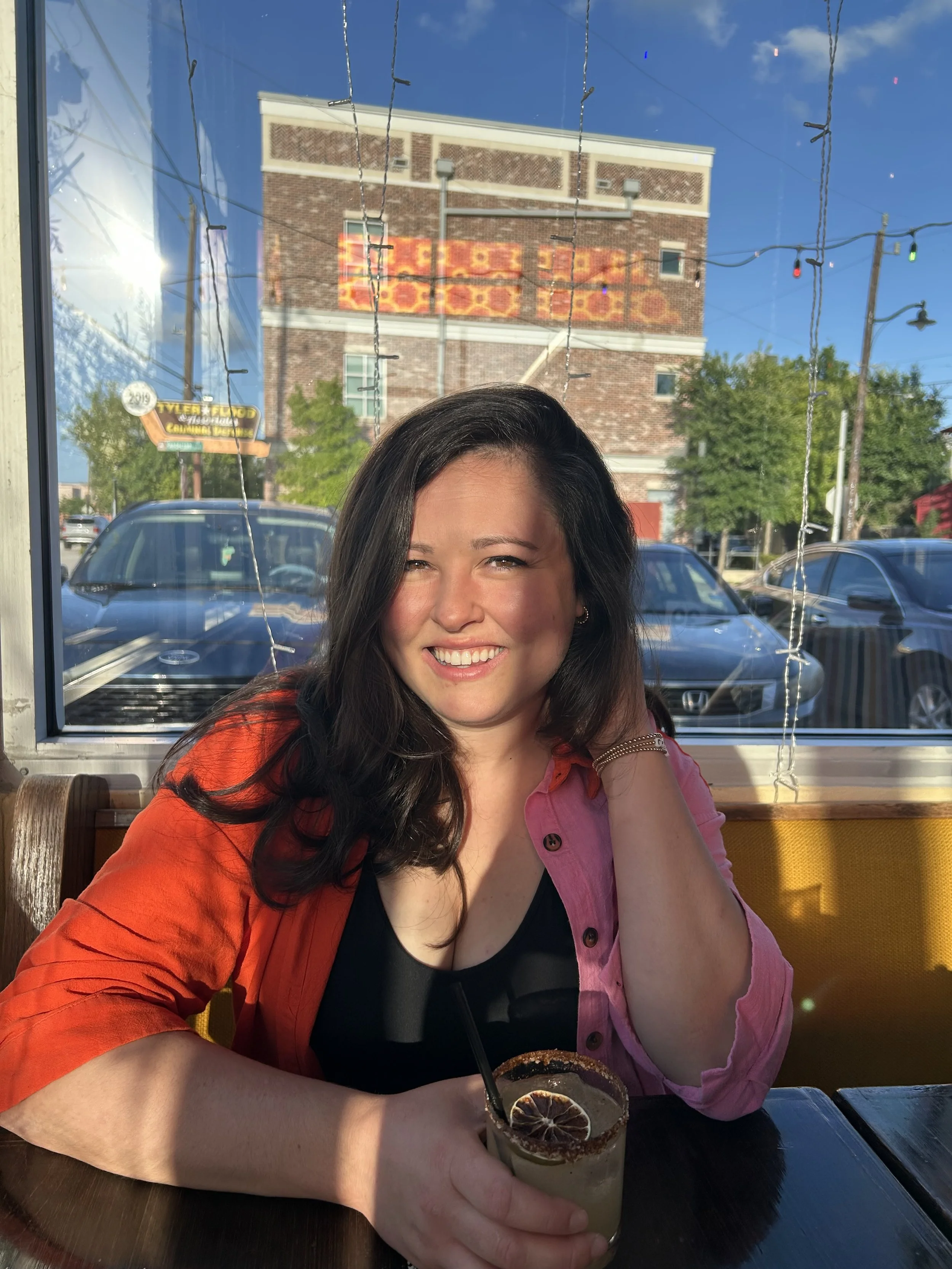 A woman with long dark hair smiling while sitting at a table in a restaurant or cafe, holding a drink with lemon and chili powder rim, with a brick and glass window reflecting a building with colored signs outside.