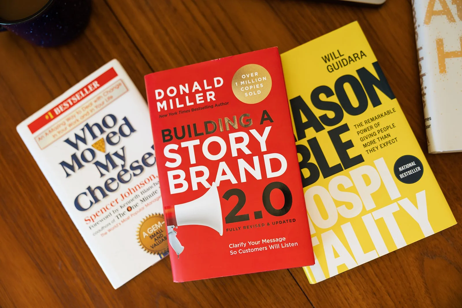 Three books on a wooden surface, including 'Who Moved My Cheese' by Spencer Johnson, 'Building a StoryBrand 2.0' by Donald Miller, and 'Able to Disappear' by Will Guidara.