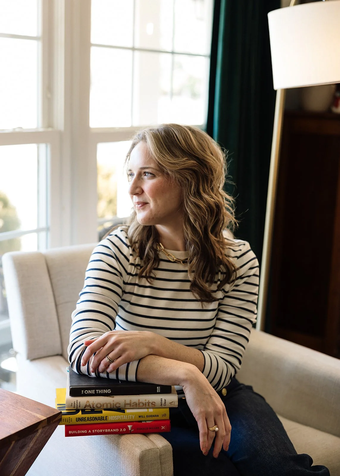 A woman with wavy brown hair and a striped shirt sitting on a cream-colored couch, holding a stack of five books, with large windows in the background letting in natural light.