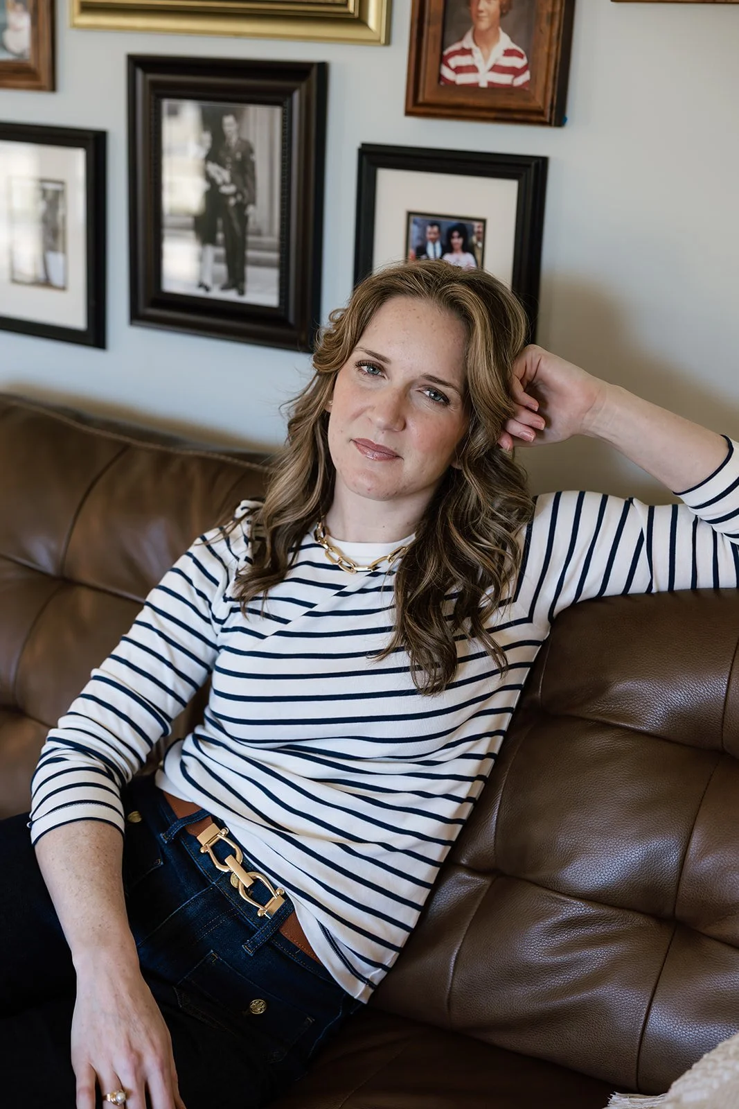 A woman with light skin and curly brown hair is sitting on a brown leather couch with her left arm resting behind her head. She is wearing a white and navy striped long-sleeve shirt, a gold chain necklace, a brown belt with a gold buckle, and dark jeans. The background shows framed photographs on a wall.