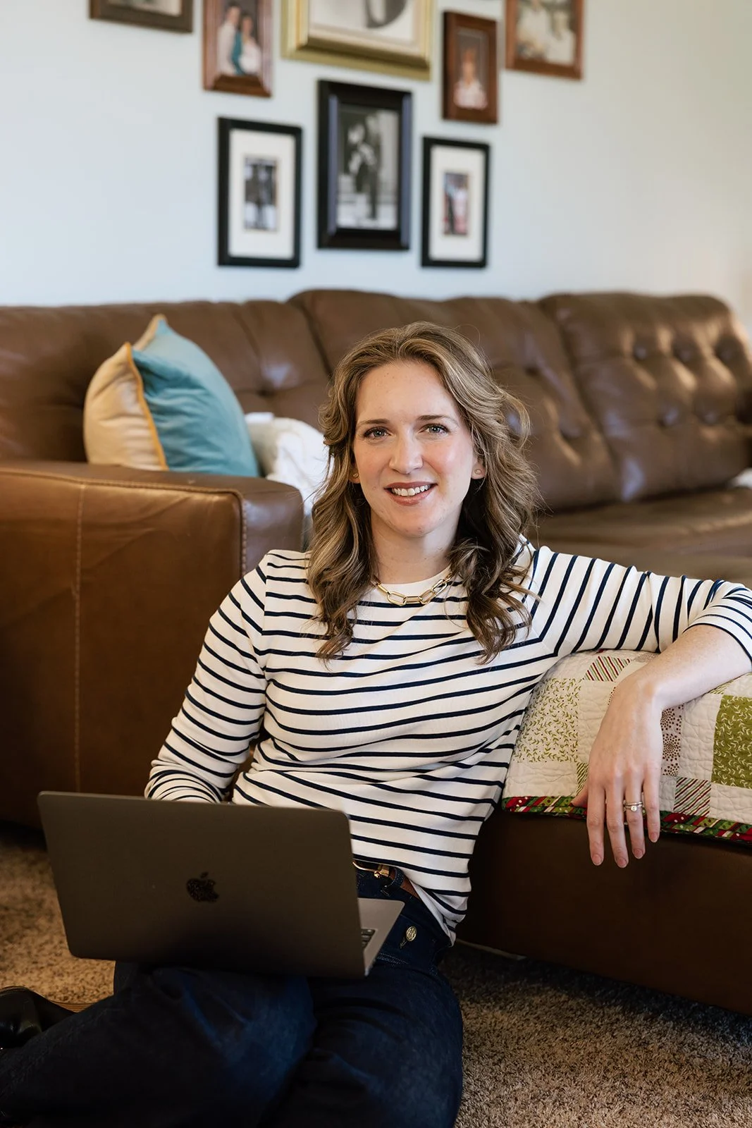 A woman with wavy hair wearing a striped shirt, sitting on the floor with a laptop on her lap, smiling, in a living room with a brown leather couch, pillows, and a gallery wall of framed pictures.