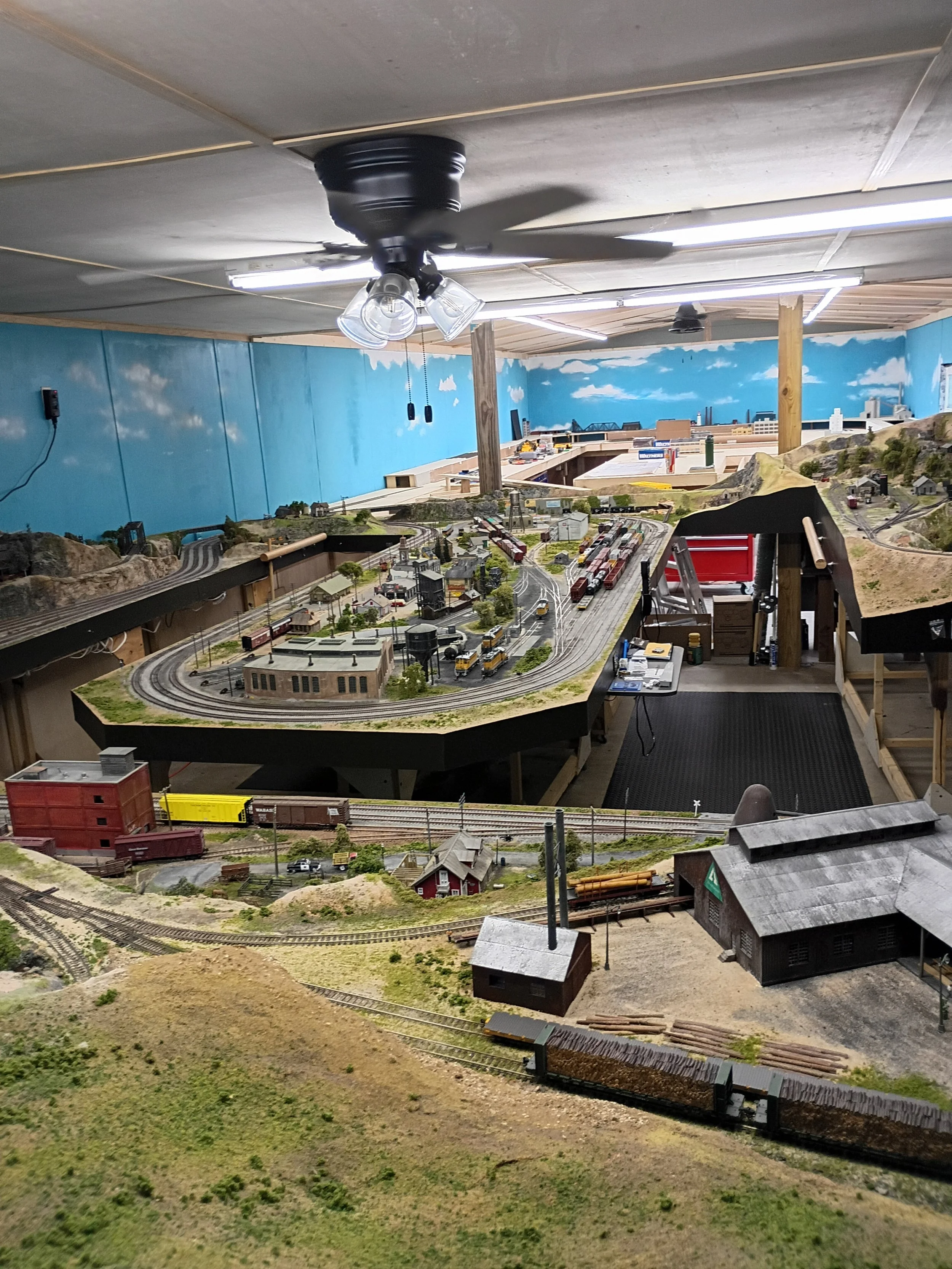 An indoor model train layout featuring tracks, buildings, trains, hills, and a painted sky mural on the back wall.