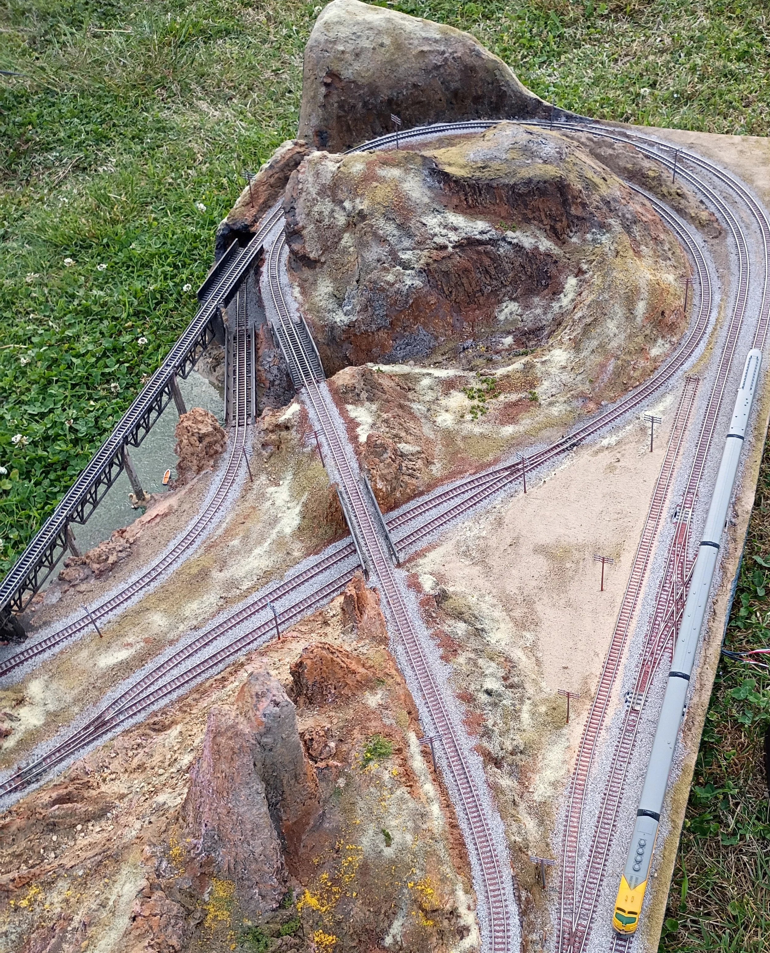A model train has tracks running over a large rock formation in an outdoor setting with grass and small plants nearby.