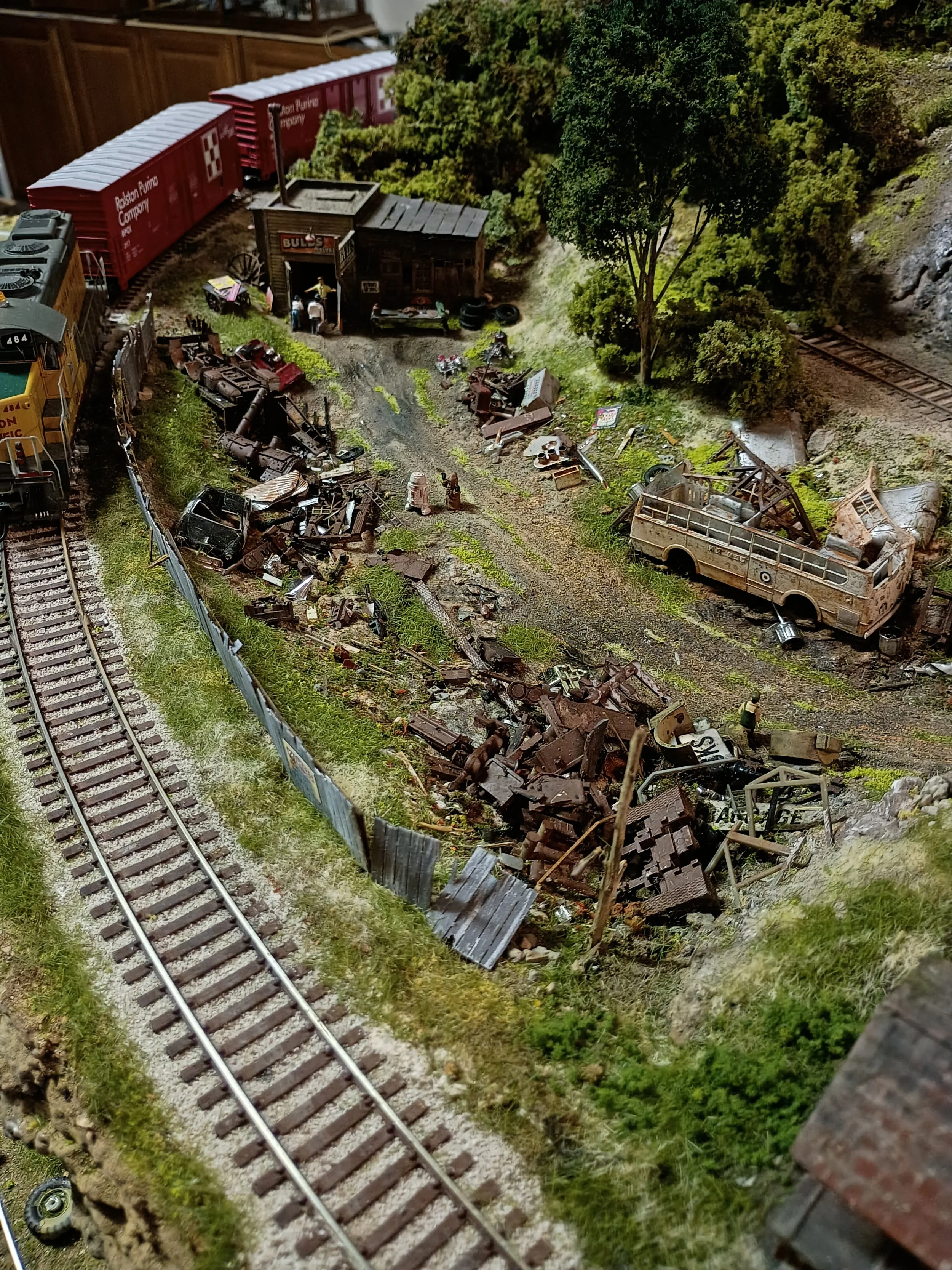 A model train scene with a damaged train, wreckage, and a small building labeled 'BULL'S' near a tree and railroad tracks.