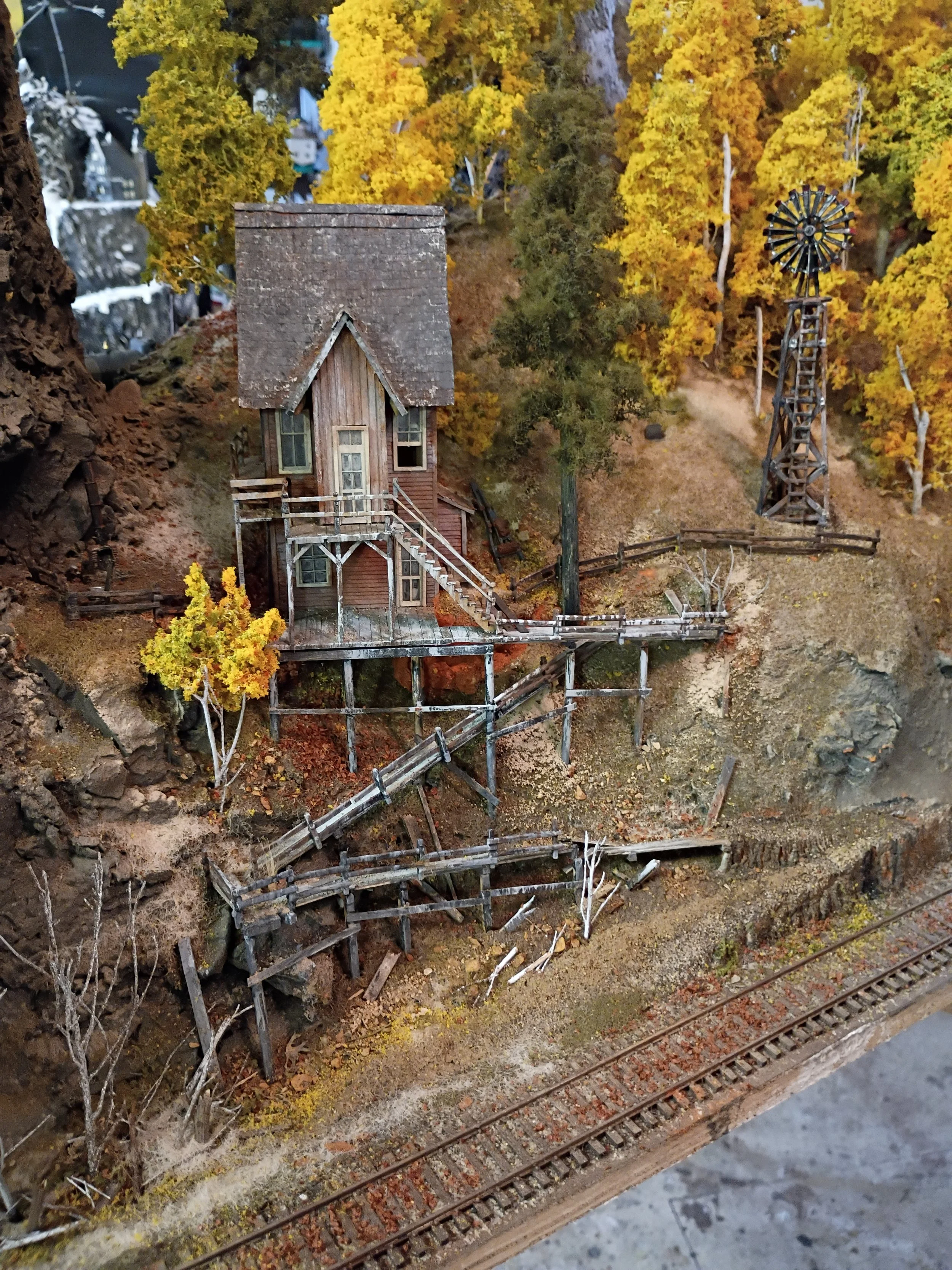A miniature model of a rustic house with a staircase and a windmill surrounded by trees and a railway track.