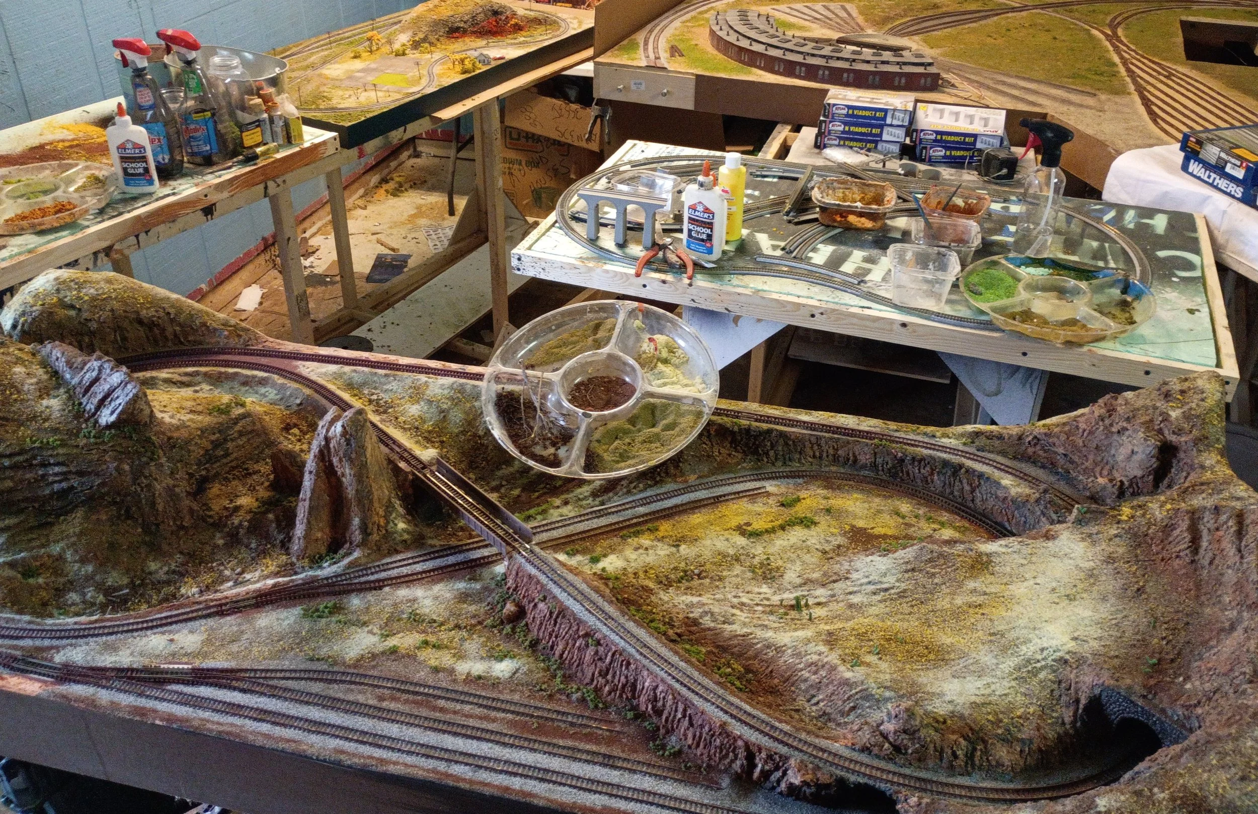 A model train setup with tracks, scenic landscape, and supplies for building and maintaining a model train layout, including glue, scenery materials, and tools.