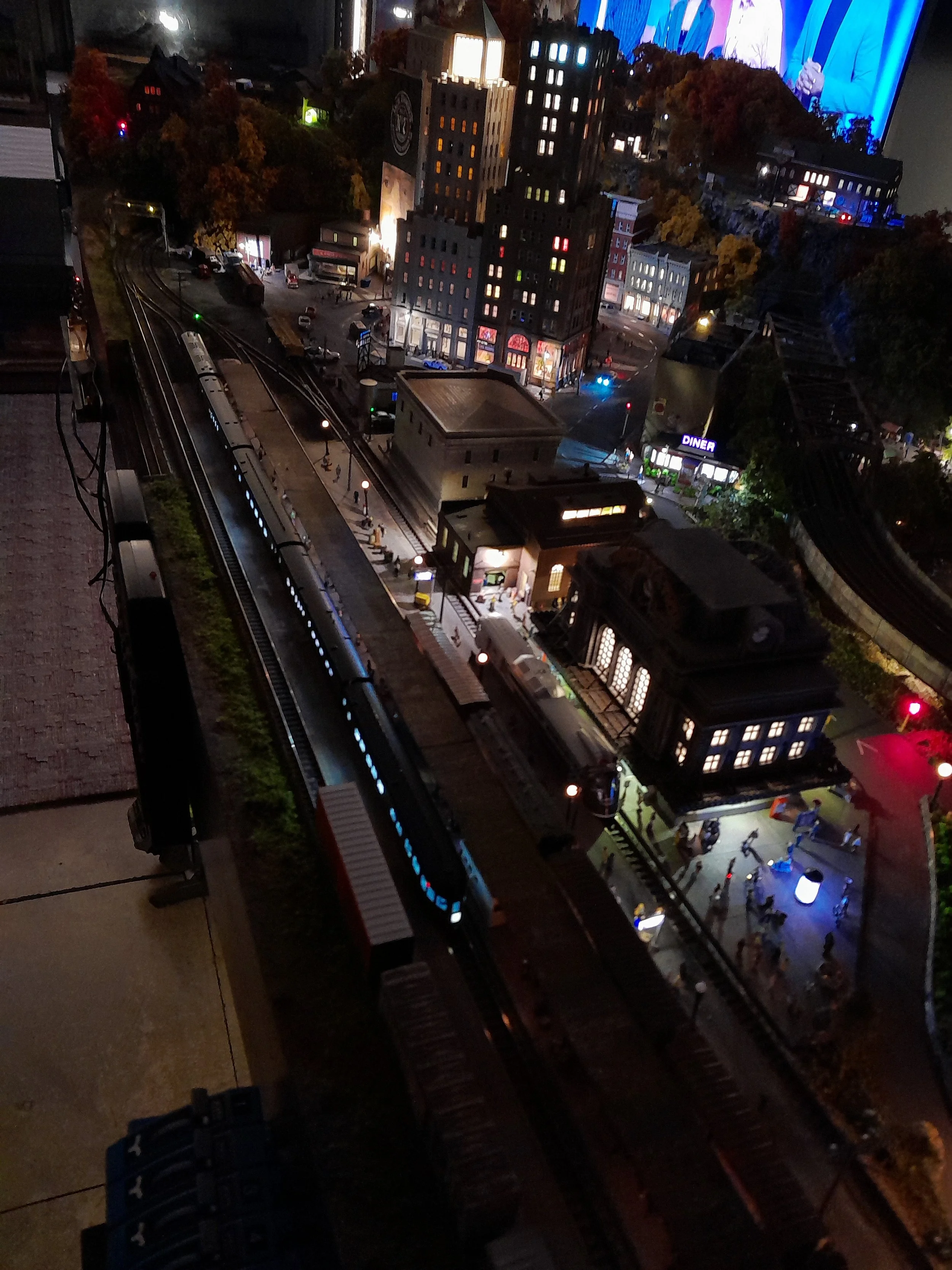 A detailed miniature model of a city street scene at night showing buildings, cars, a train track, and streetlights, with a digital screen in the background.