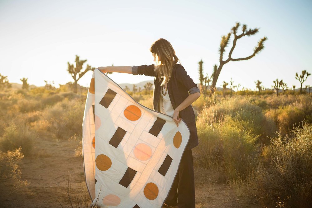 woman flapping white cotton quilt with charcoal rectangles and orange and pink circles