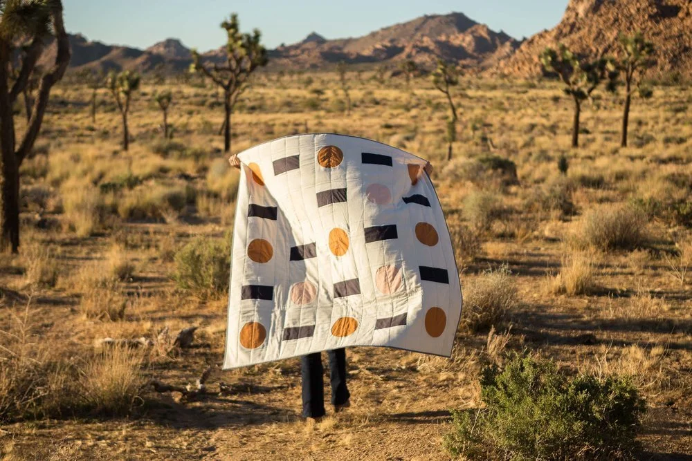 woman holding white cotton quilt with charcoal rectangles and orange and pink circles in desert