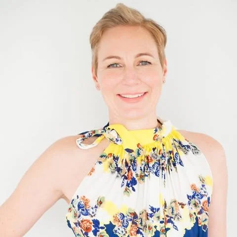 Portrait of a smiling woman with short blonde hair wearing a sleeveless floral dress with a gathered neckline.