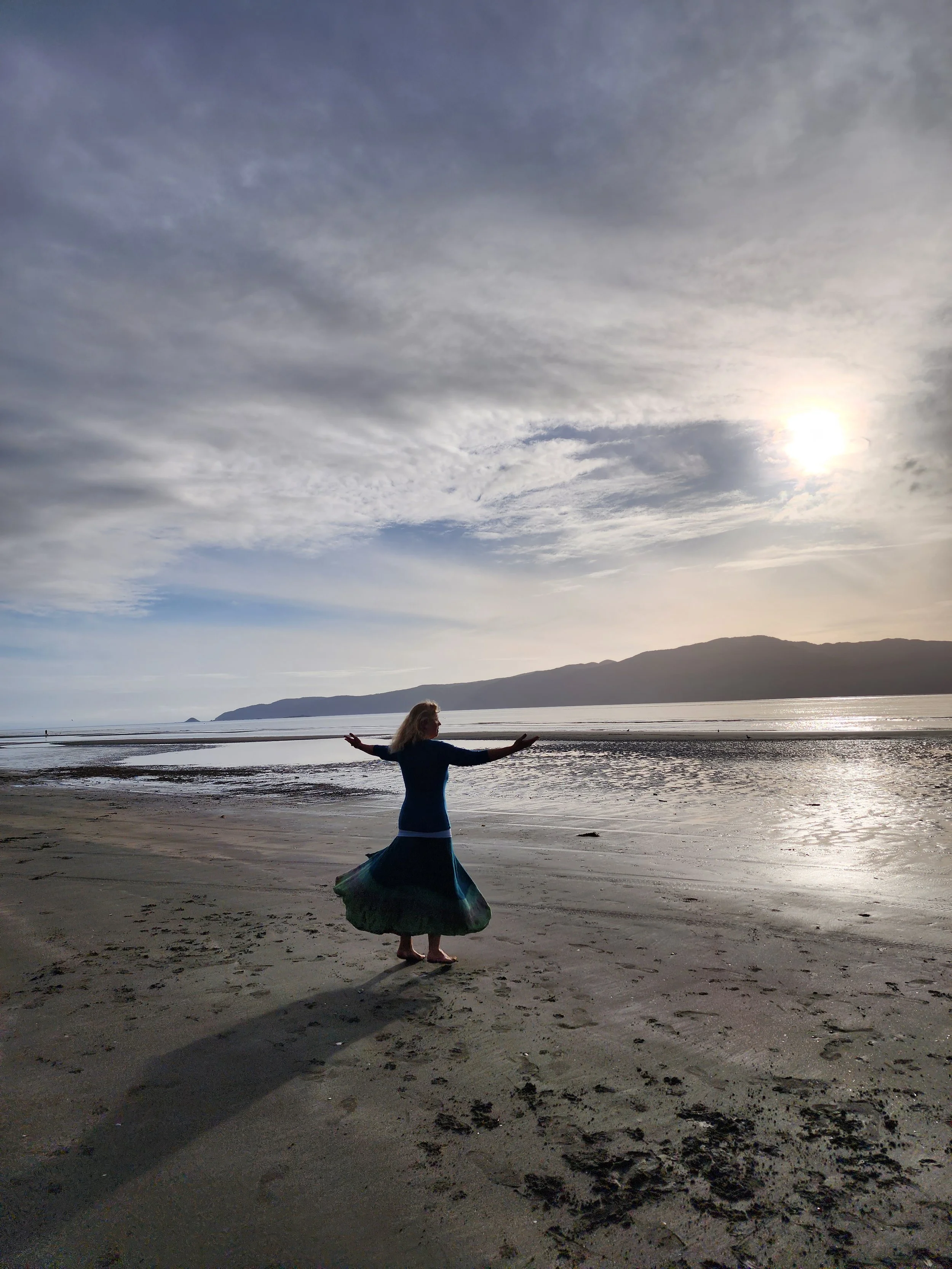 A woman in a long skirt standing on a sandy beach with her arms extended, facing the ocean, during sunset or sunrise, with hills in the background and clouds in the sky.