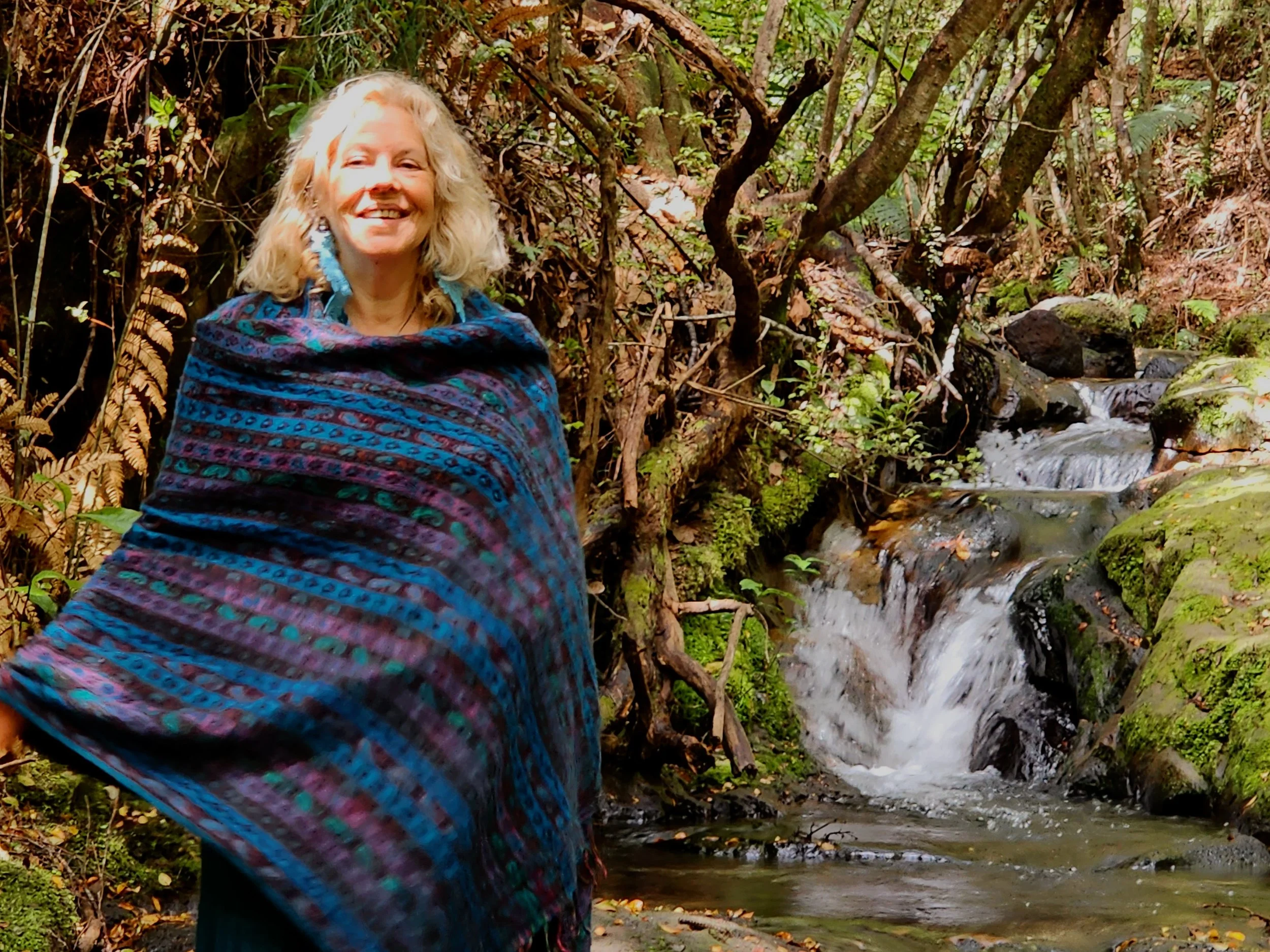 Woman with curly blonde hair smiling outdoors near a small waterfall in a forest, wrapped in a colorful, patterned shawl.