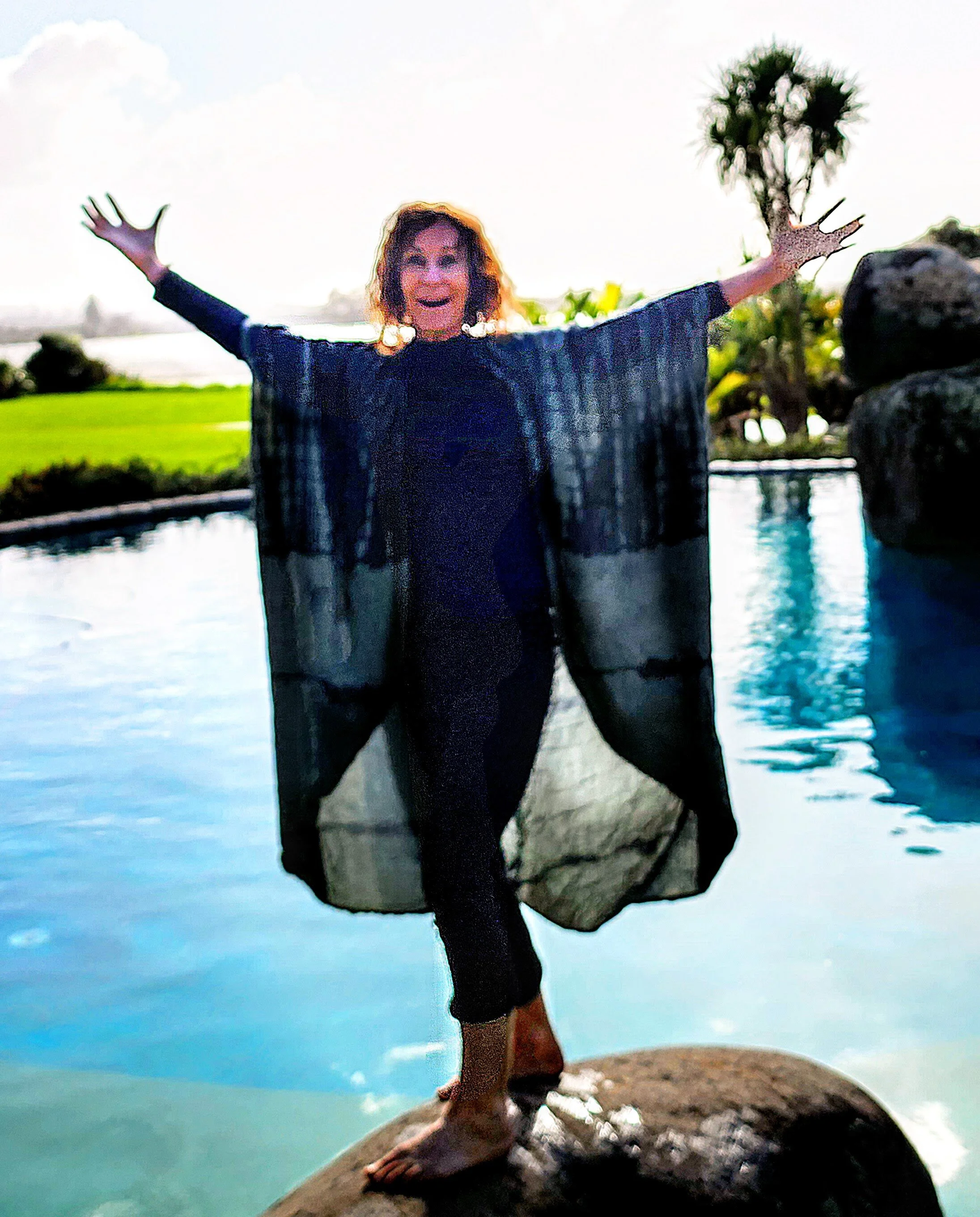 Woman standing on a rock with arms outstretched, by a pool with water features and green landscape in the background.