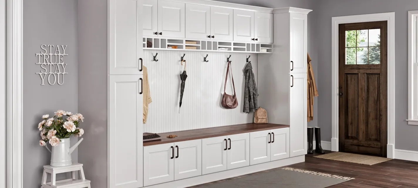 A mudroom with white cabinets, a wooden bench, hooks with hanging jackets and accessories, a flower arrangement in a white pitcher, and a brown wooden door.