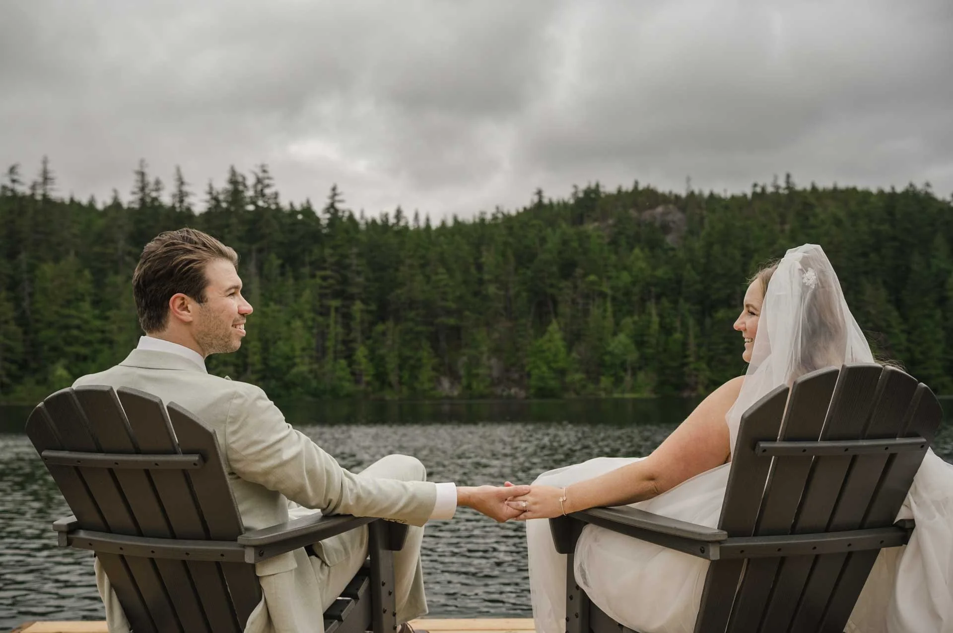 bride and groom sitting on muskoka chairs by the lake with mountains in the background